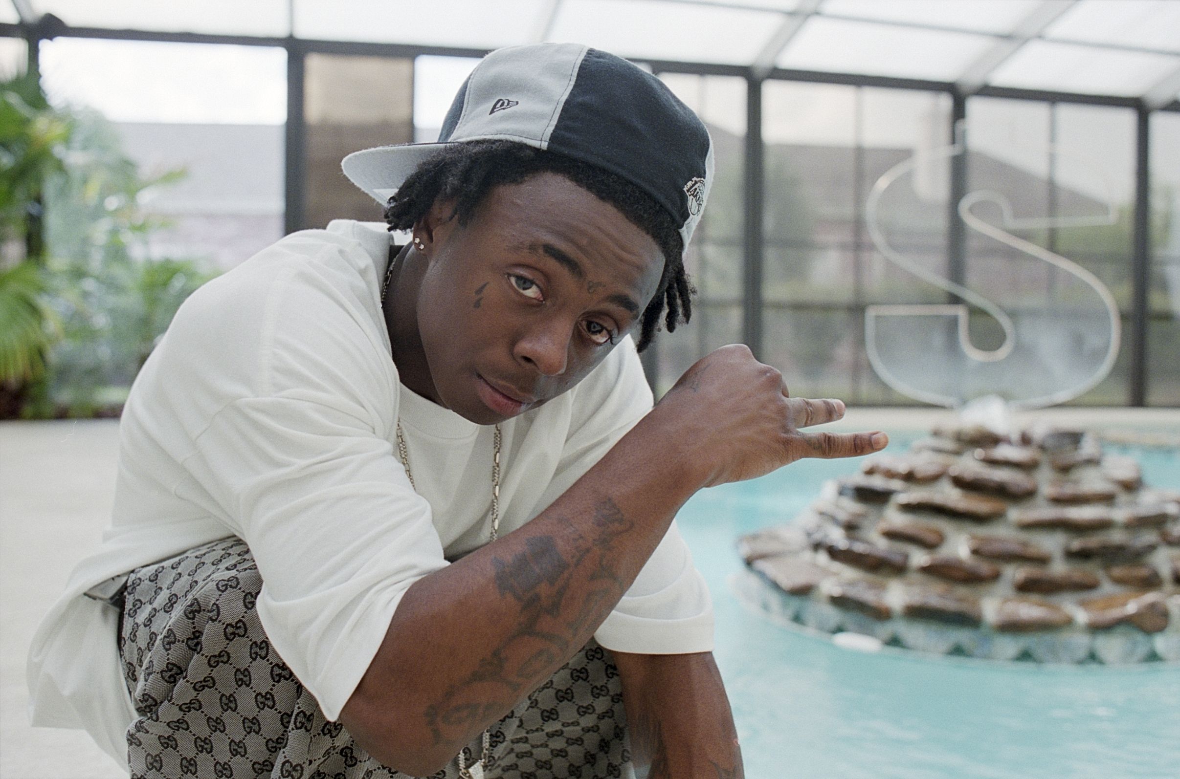 Lil Wayne crouches next to an indoor swimming pool, in the center of which is a fountain with a clear acrylic dollar sign symbol.