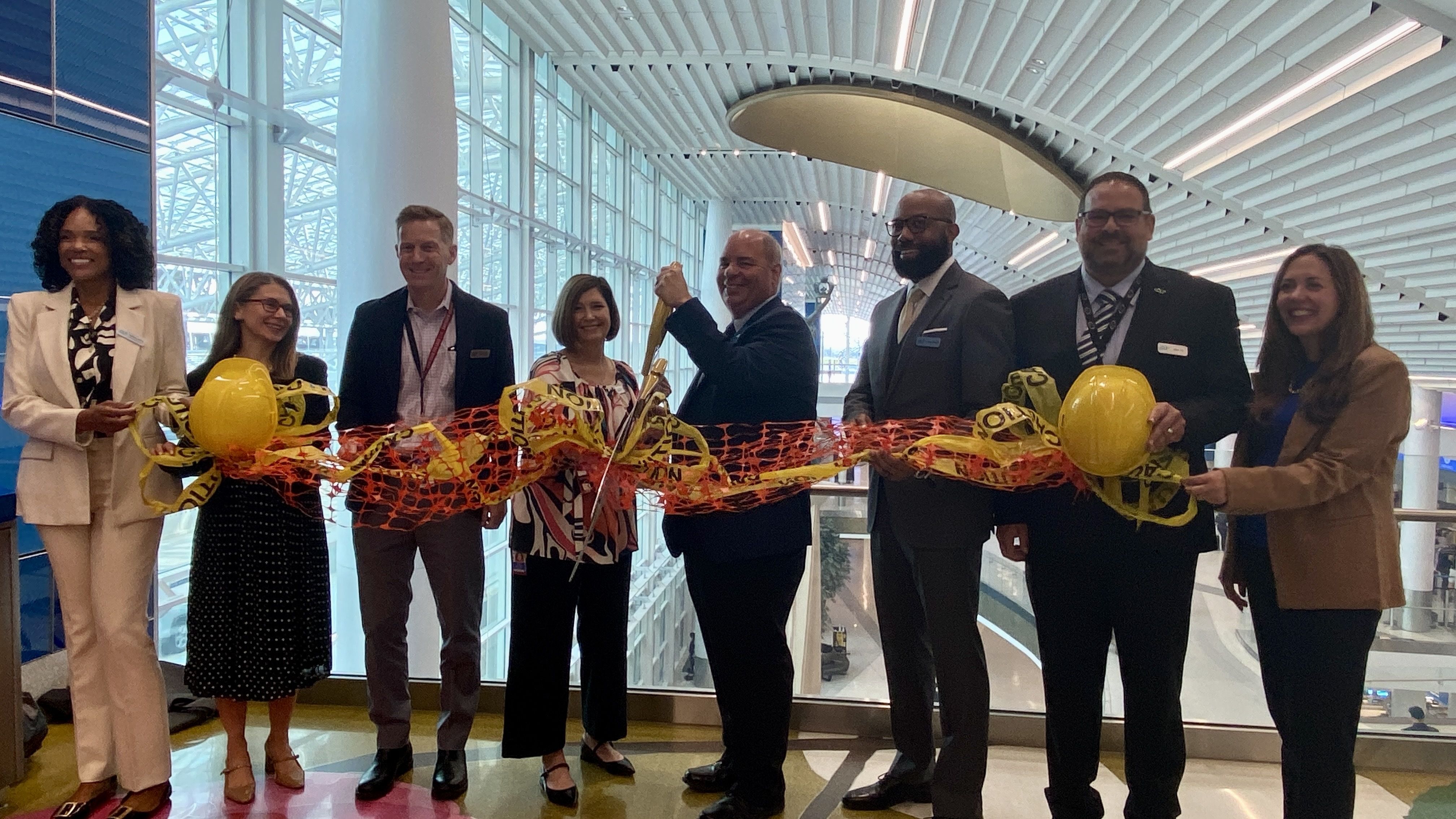 Eight people in business attire stand indoors holding a long orange construction netting with yellow caution tape and two yellow hard hats, one man in center cutting the ribbon with scissors.