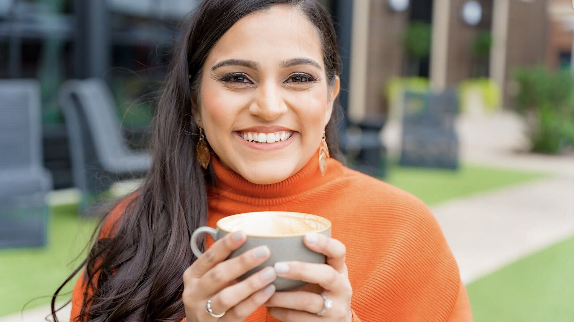 A woman wearing an orange sweater, drinking a cup of coffee