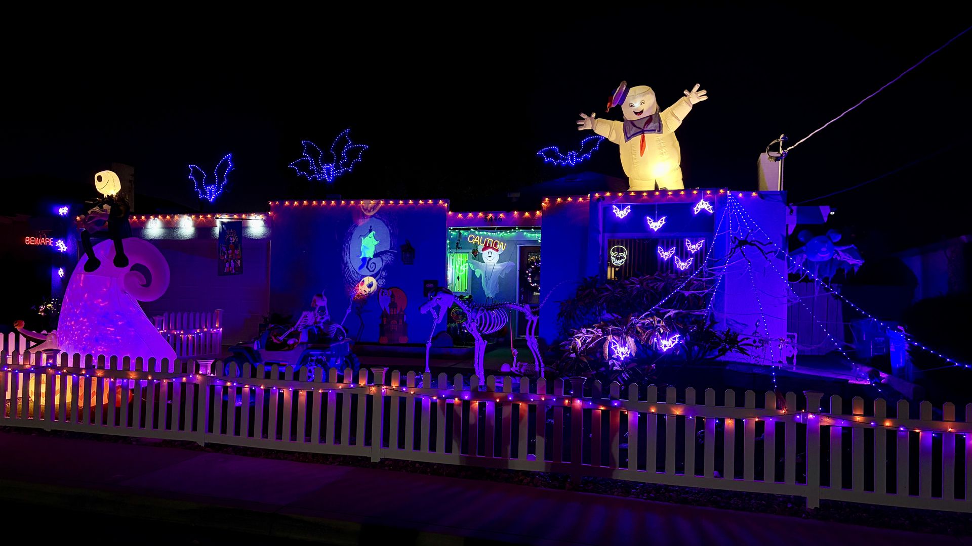 Night scene of a Halloween parade float featuring a large yellow inflatable figure with arms raised, blue and purple lights, neon bats, a white picket fence, skeleton decorations, and a CAUTION sign.