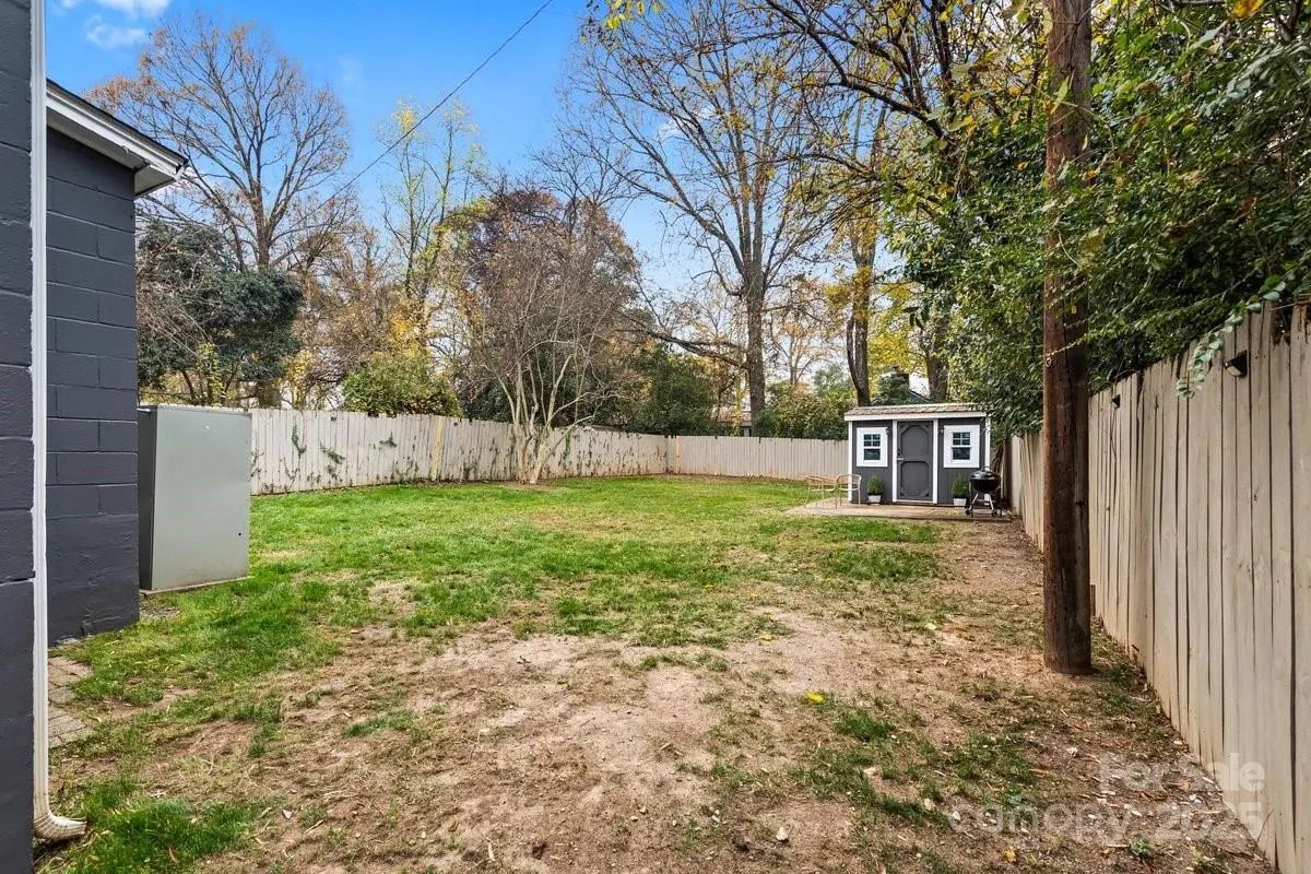 Backyard with patchy grass, a small gray garden shed with white trim, wooden fence, leafless trees, and a blue sky with scattered clouds.
