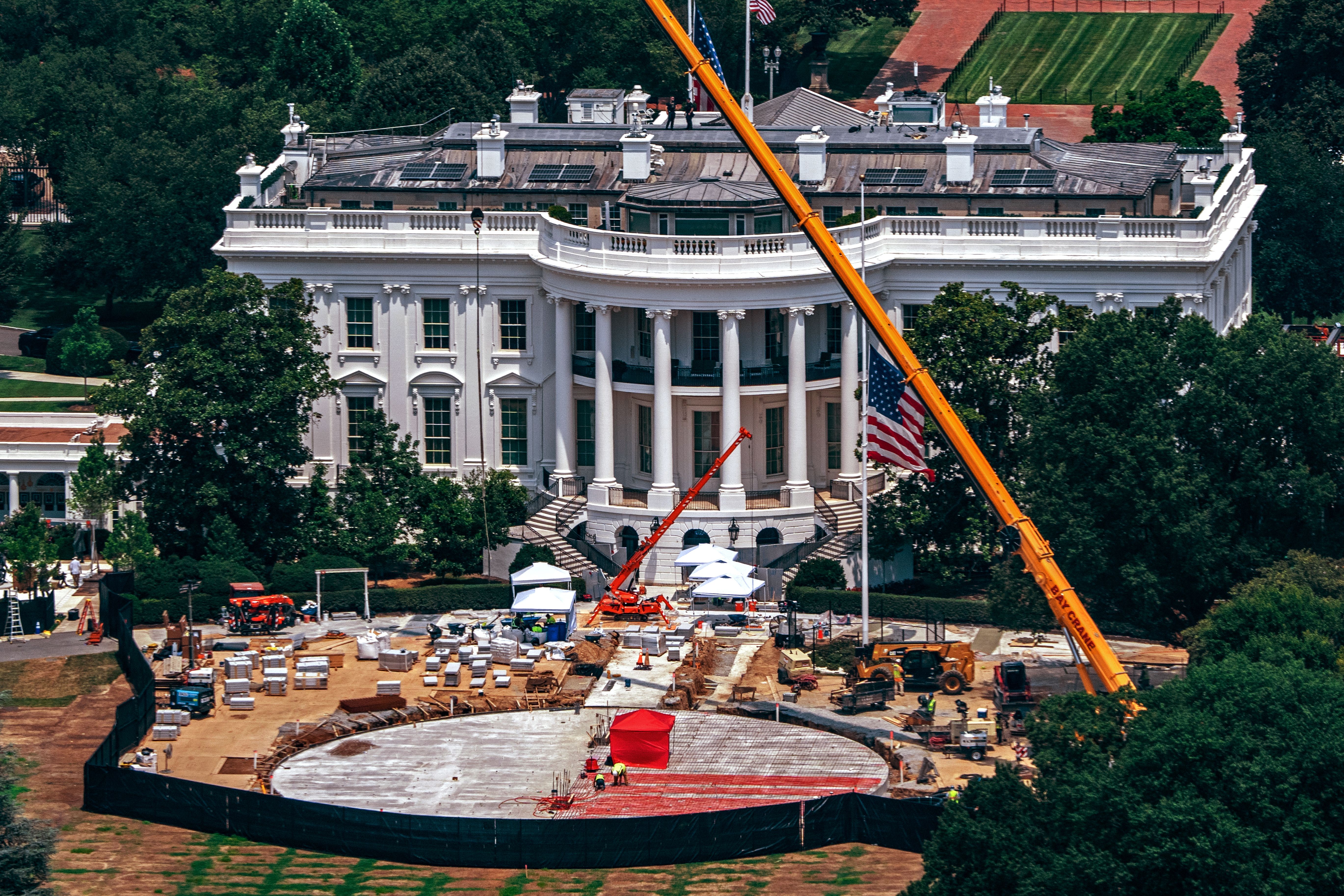 Aerial view of the White House under construction, with a large orange crane across the front yard, workers and equipment on the circular forecourt, surrounded by trees.