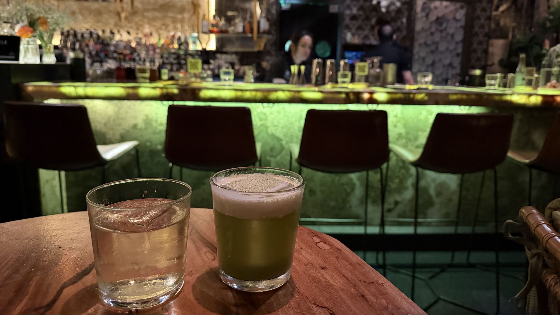 Two drinks on a wooden table in front of a dimly lit bar with green under-counter lighting and several empty stools, with bartenders and bottles in the background.
