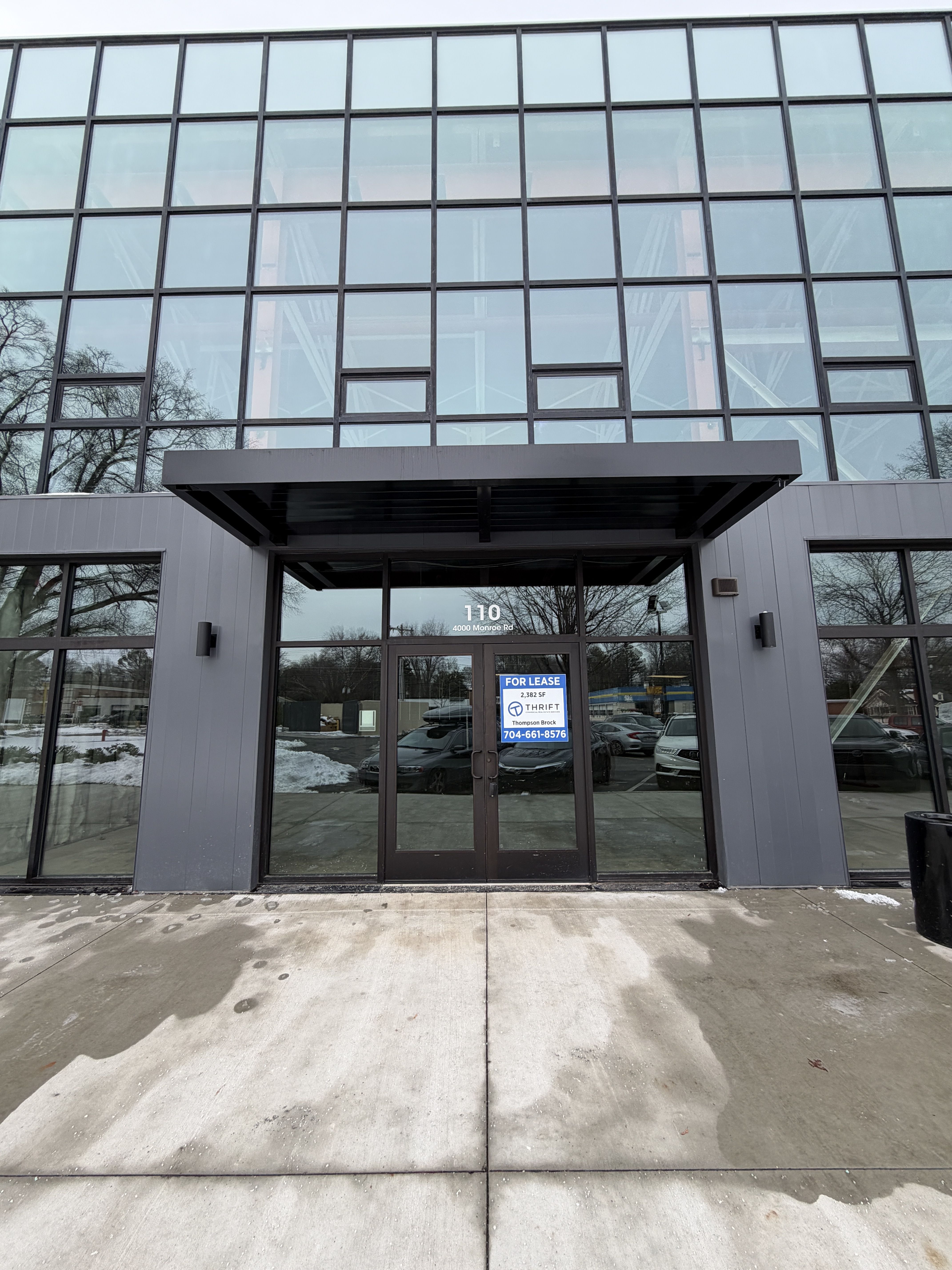 Gray modern commercial building with large glass windows and a black framed glass door displaying a "For Lease" sign with contact info, address 110 4000 Monroe Rd visible above the door.