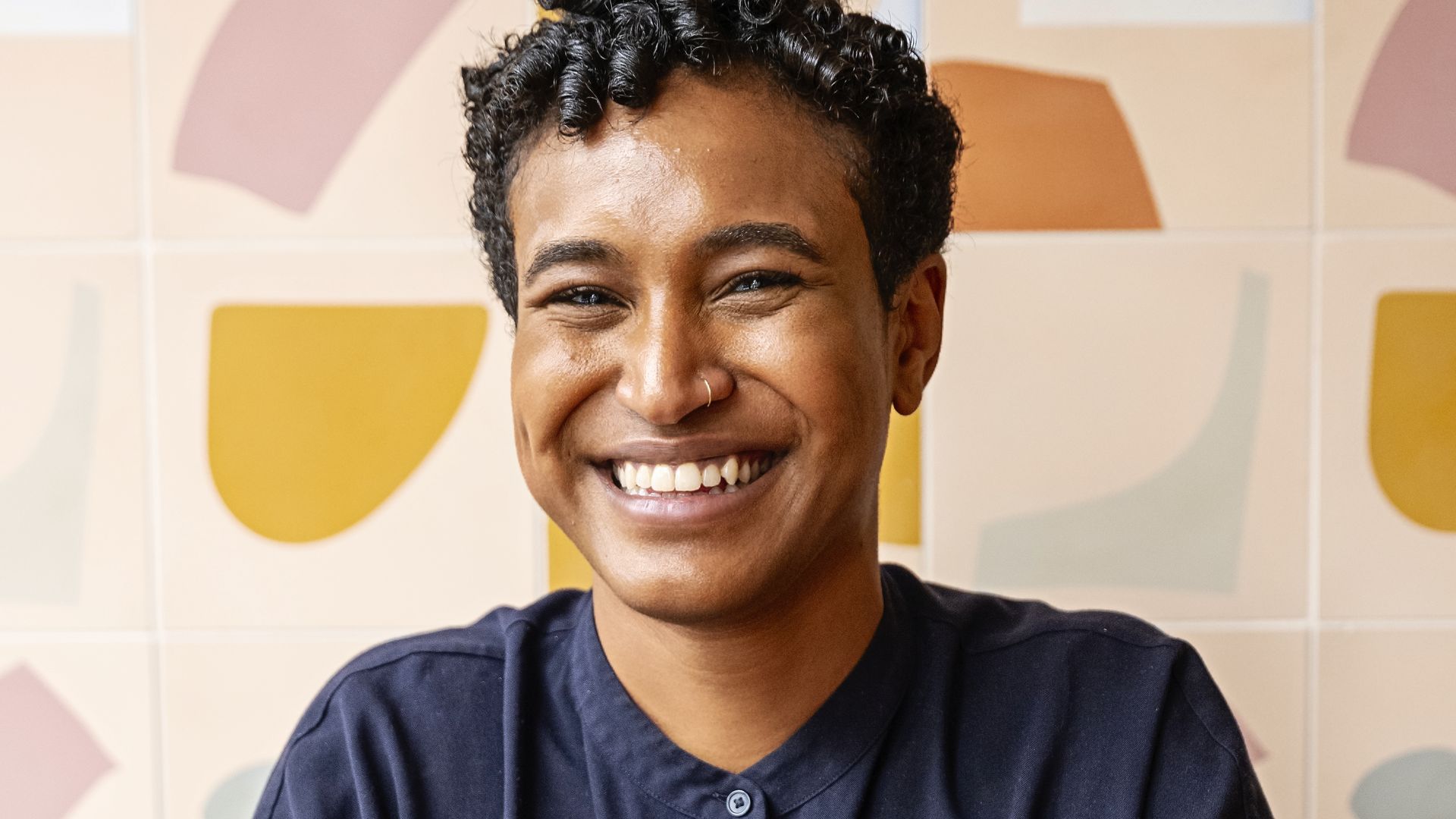 A person with short curly hair wearing a short-sleeve button-down shirt smiling big. She has her hands folded in front of her and is sitting in front of a wall with modern, abstract shapes on it.