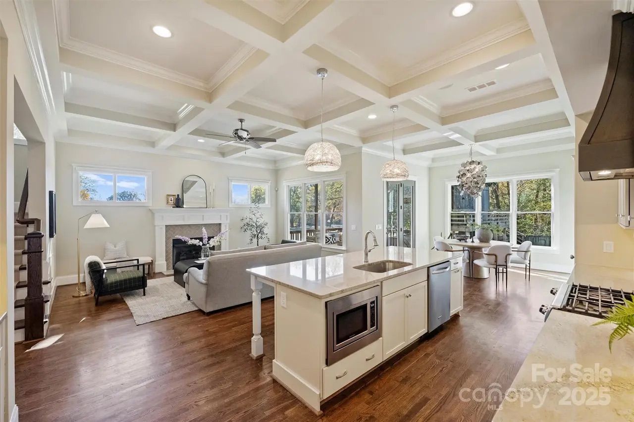 Bright open-concept living room and kitchen with white coffered ceiling, hardwood floors, large island with sink, stainless steel appliances, fireplace, and light-filled dining area with large windows.