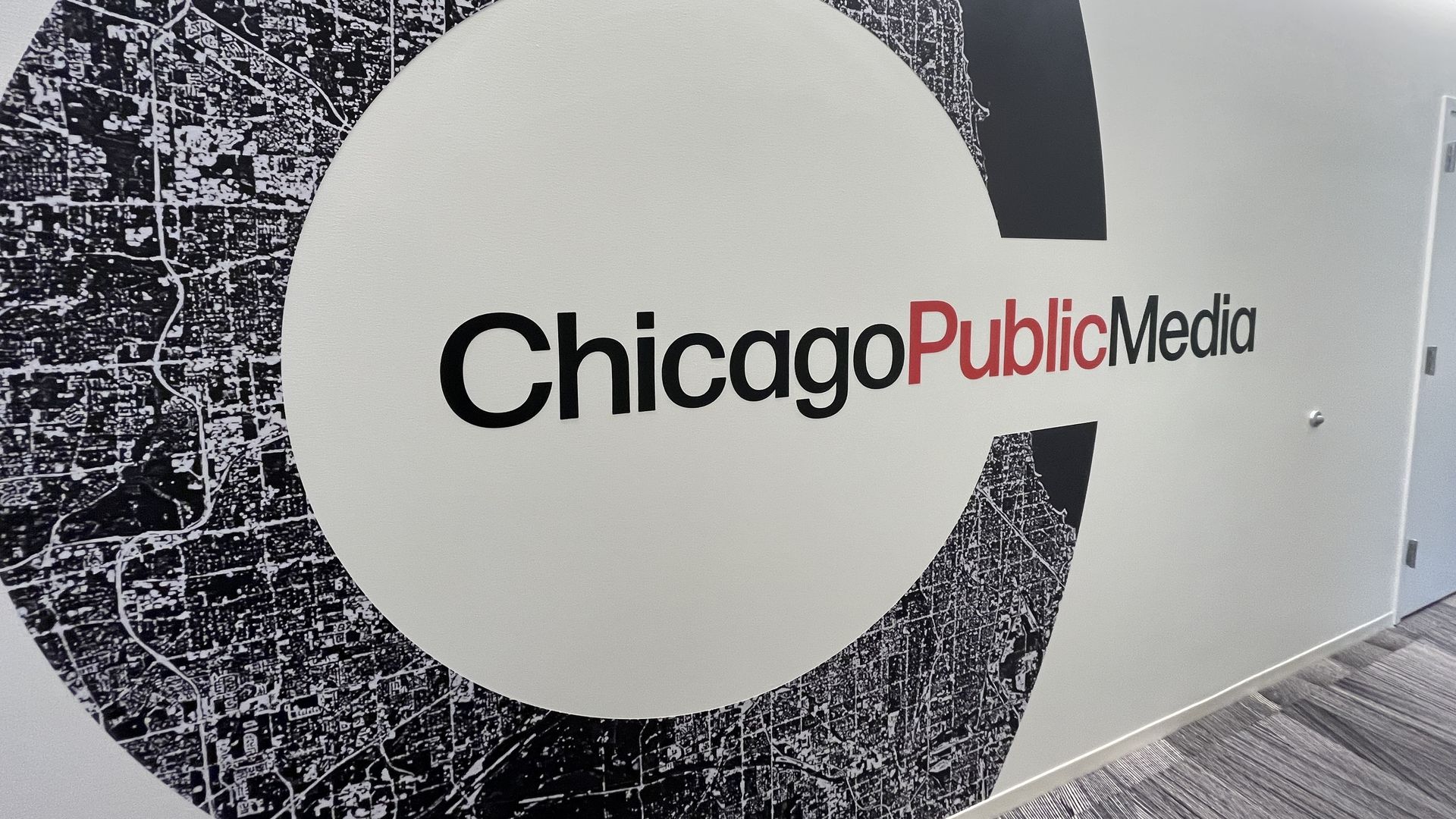 Wall with large black and white stylized 'C' map design and the words Chicago Public Media, with Public in red and others in black, in an office setting with gray carpet.