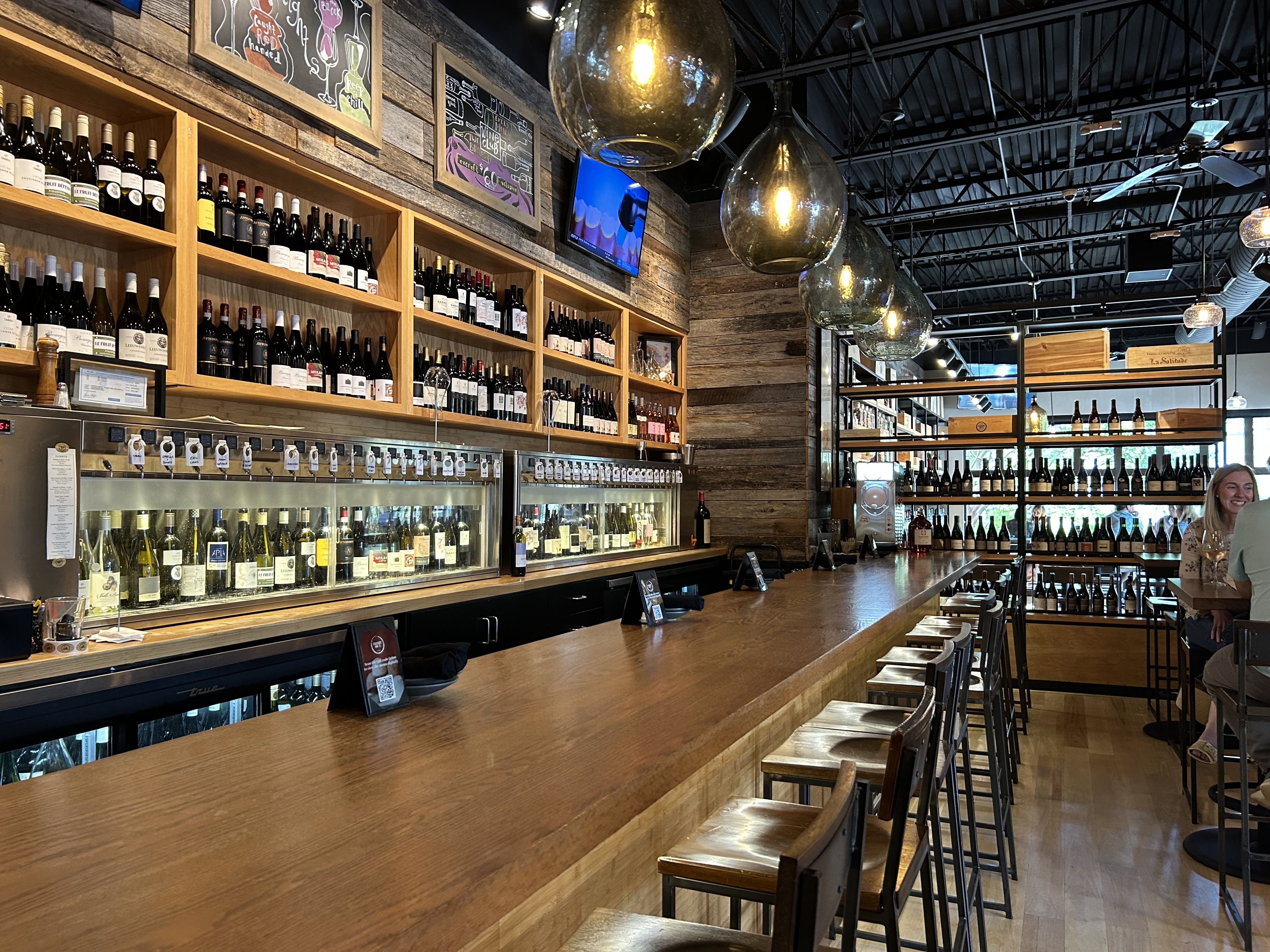 Interior of a modern wine bar with wooden countertops, shelves stocked with wine bottles, hanging glass pendant lights, and people enjoying drinks at tables.