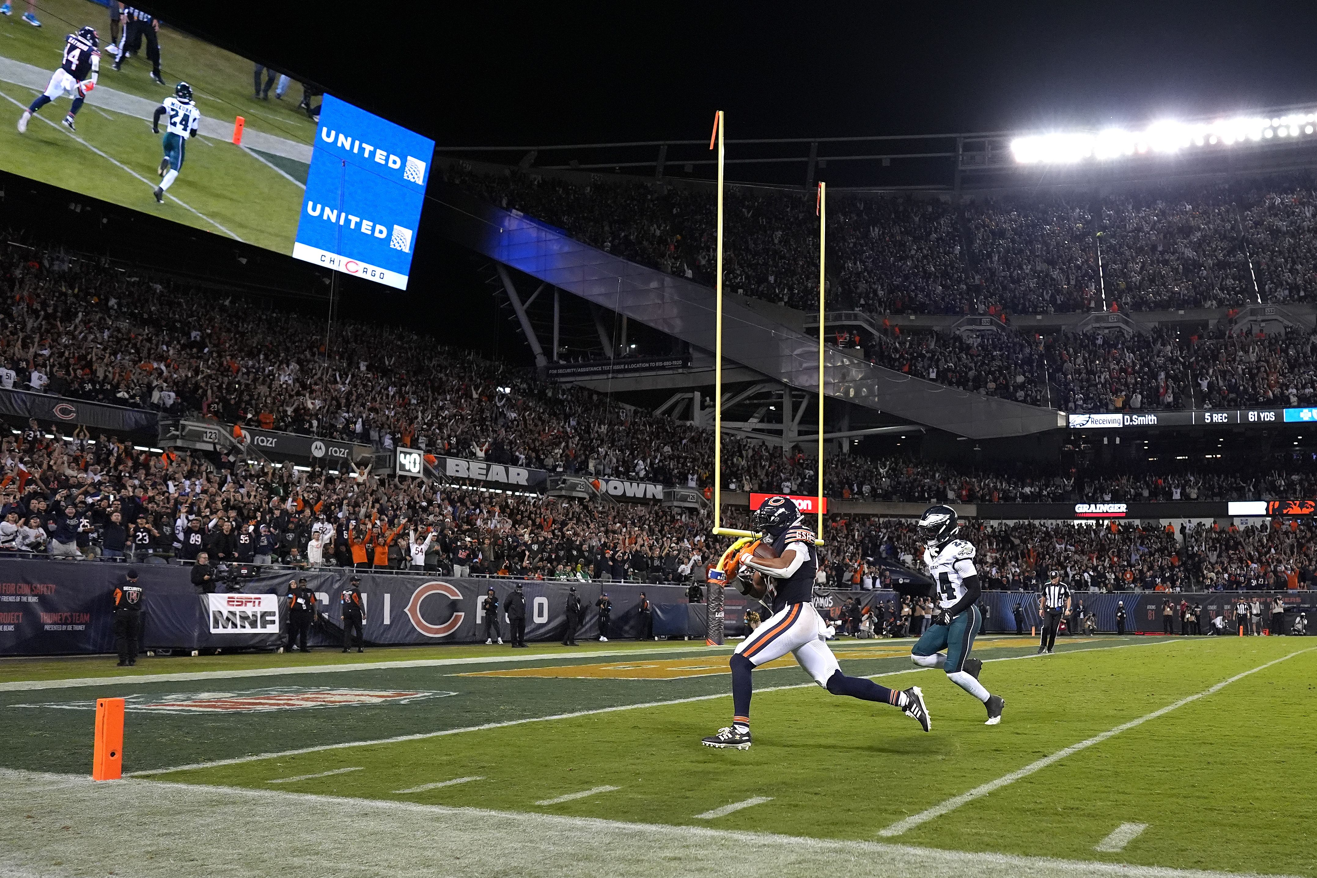 Night football game in a packed stadium. Two players near end zone - one in white, one in dark - chase a play as a large screen shows "UNITED" and Chicago branding. Bright lights, yellow goalposts.