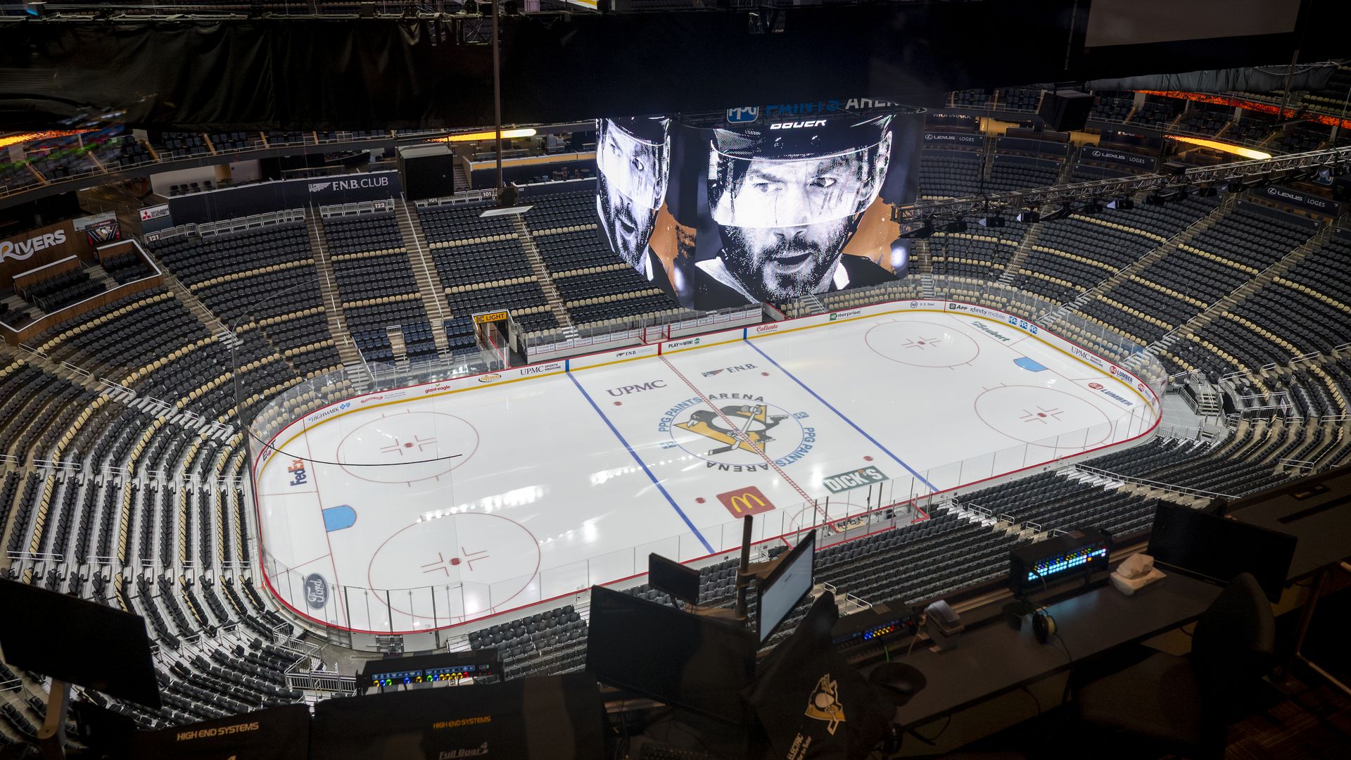 Indoor hockey arena with empty dark seats surrounding a brightly lit ice rink. A large hanging screen shows a masked hockey player; Penguins logo at center ice.