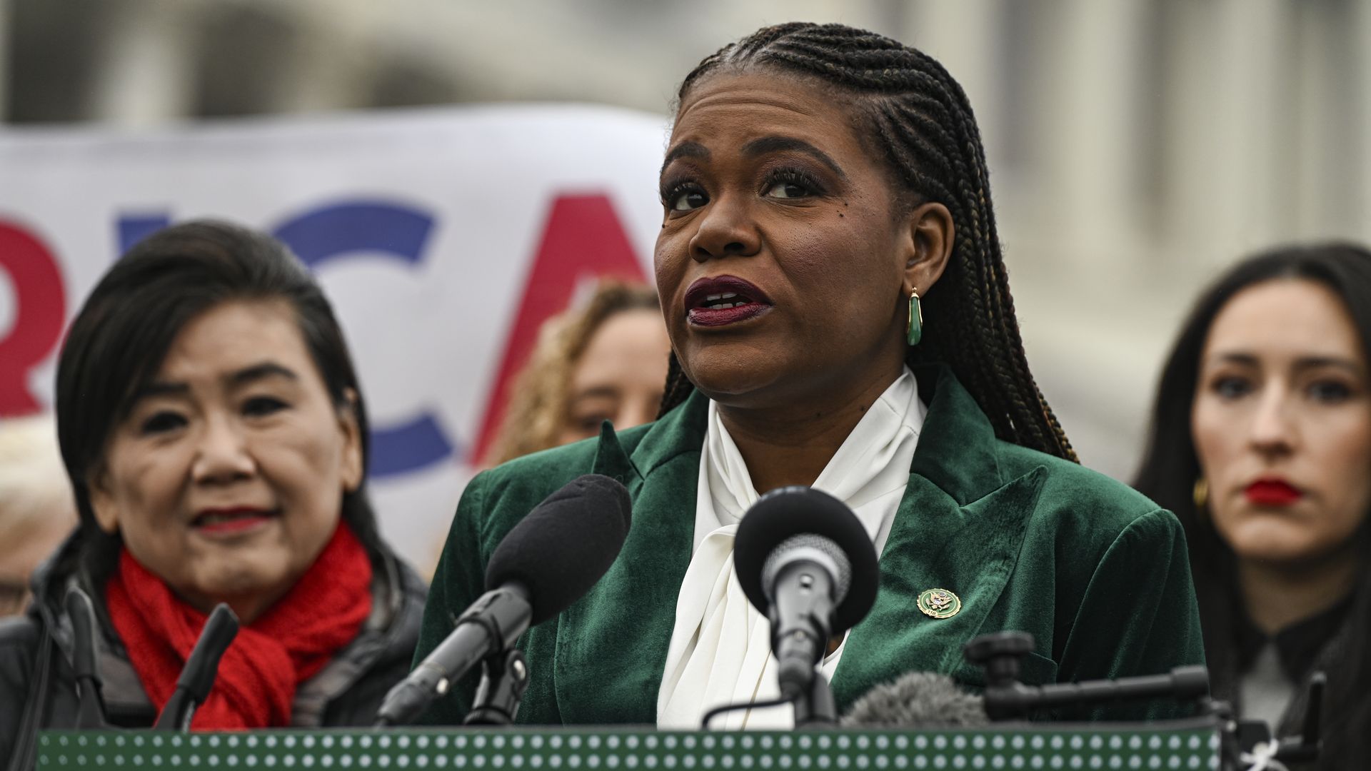 Black woman with braided hair in a green blazer and white blouse speaks at a press conference, microphones in front, with others and a blurred banner in the background.