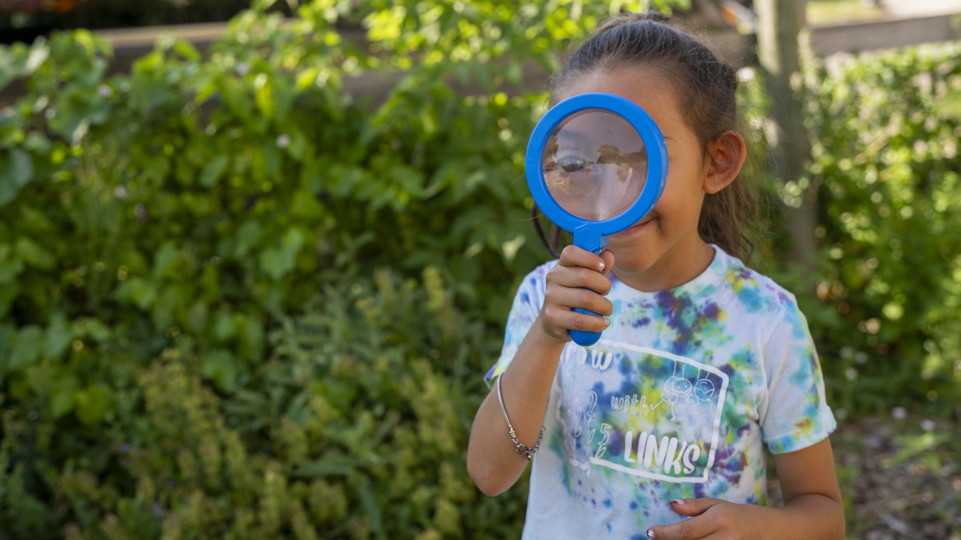 A girl in a colorful tie-dye shirt holds a blue magnifying glass up to her face, enlarging one eye, standing outside near green bushes in sunlight.