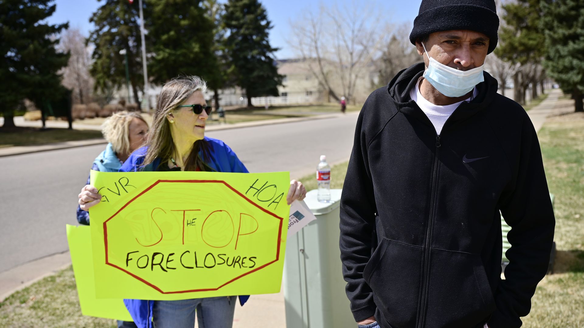 A woman stands with a bright sign that says STOP FORECLOSURES while a man stands in the foreground. 