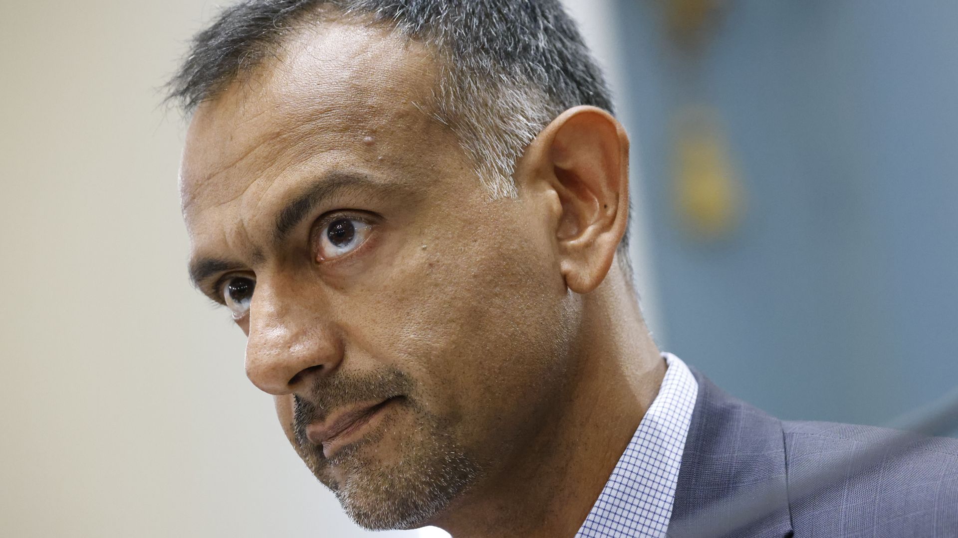 the head of a man looking upward, with short-cropped hair, a blue-grey suit jacket and checked white shirt. 