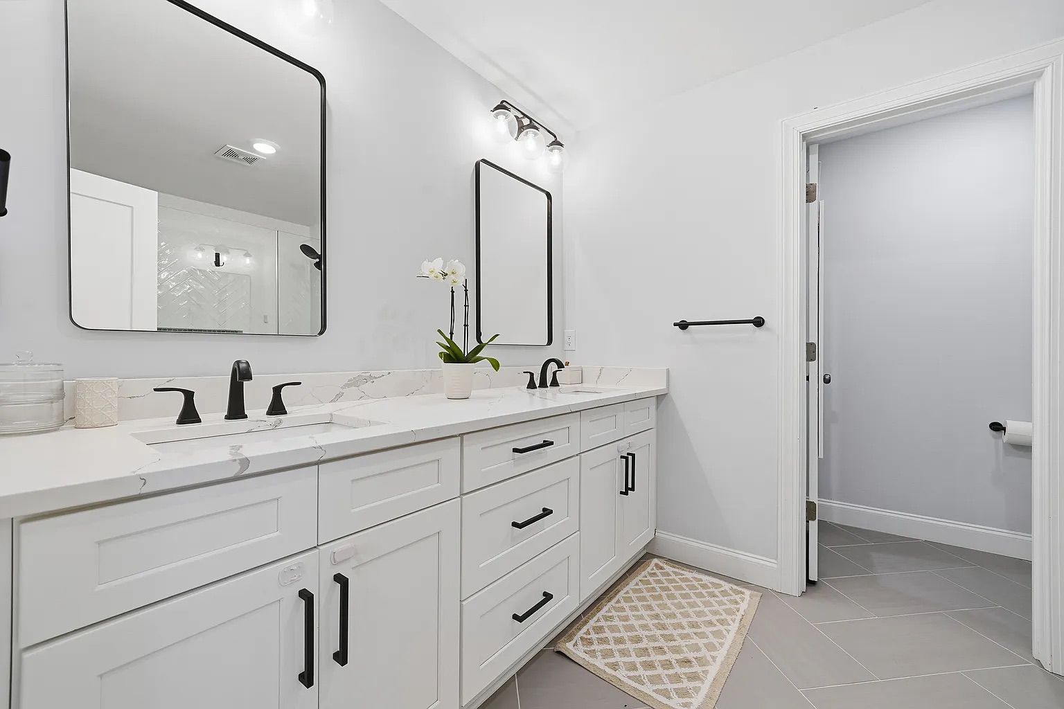 Modern white bathroom with double sink vanity, black fixtures, two black-framed mirrors, white orchid in pot, beige patterned rug, and gray tiled floor, leading to a toilet area.