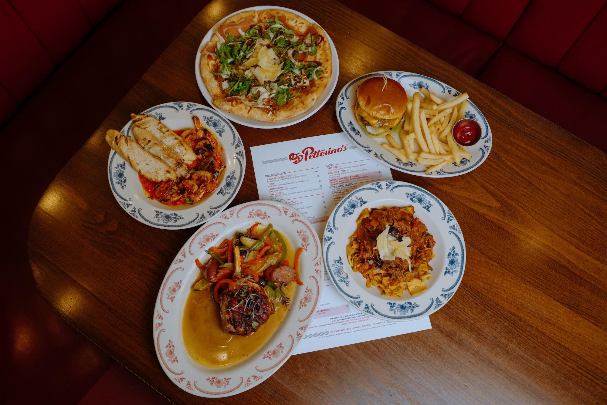 Top-down view of a wooden table with several plates of food: a greens-topped pizza, a hamburger with fries, a dish in tomato sauce with bread, and two pasta dishes; a menu sits in the center.