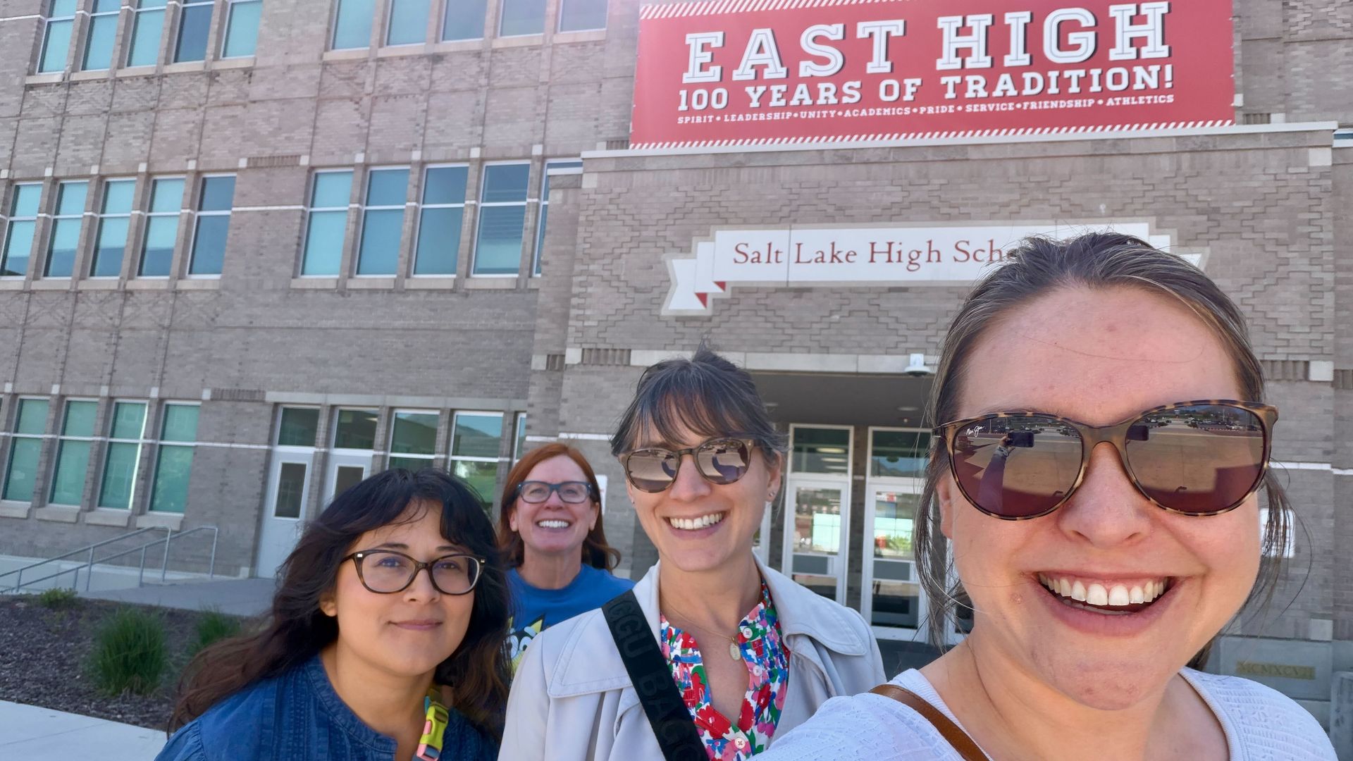 Four smiling women taking a selfie outside a brick high school. A large red banner reads "EAST HIGH 100 YEARS OF TRADITION!" and a nearby sign says "Salt Lake High School."