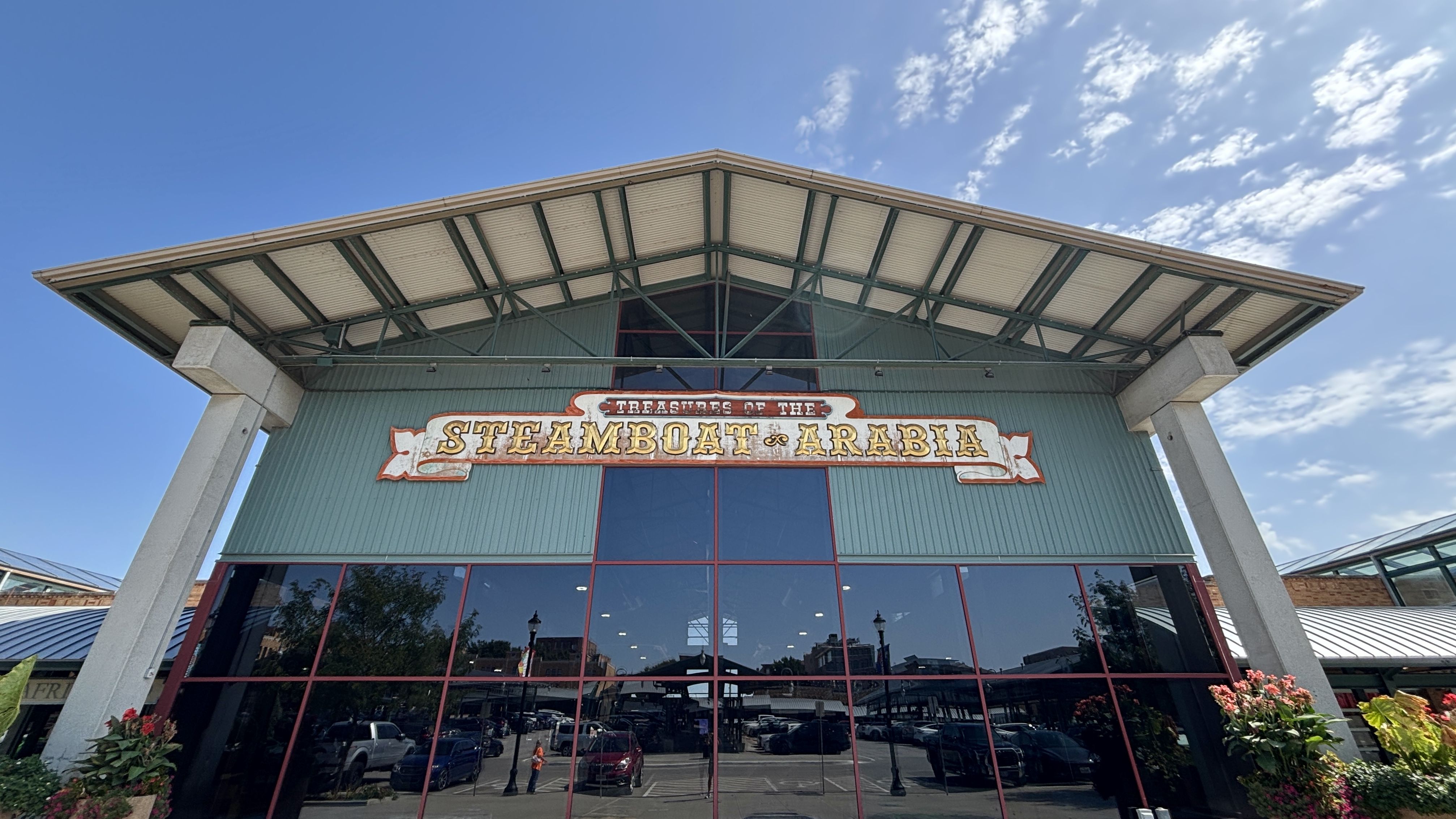 Low-angle view of a teal museum building with a metal roof and large glass façade; a decorative sign reads "STEAMBOAT ARABIA" with "TREASURES OF THE" above. Bright blue sky with scattered clouds.
