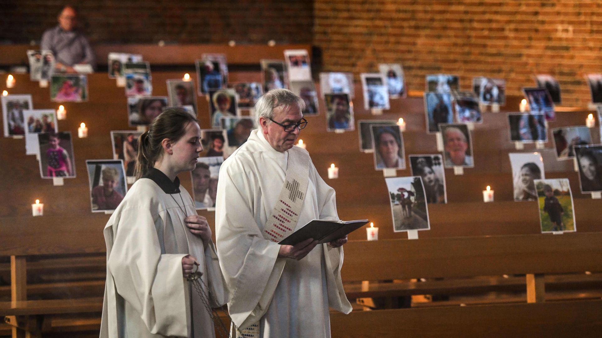 In this image, two pastors read scripture in front of benches with pictures of people