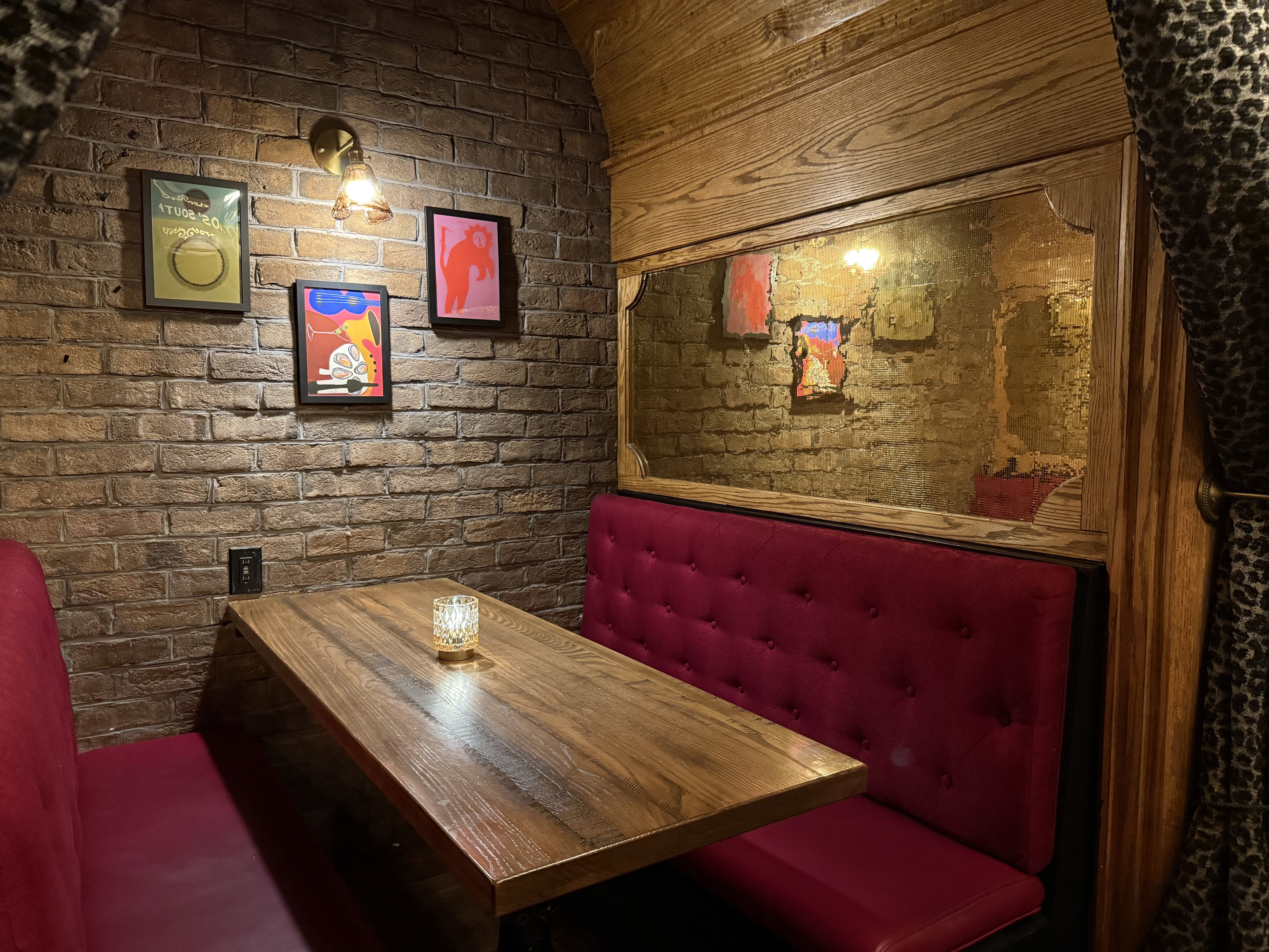 A cozy restaurant corner with a brick wall, framed colorful posters, a brass wall lamp, a wooden table, and red tufted booth seating beside a large wooden-framed mirror.