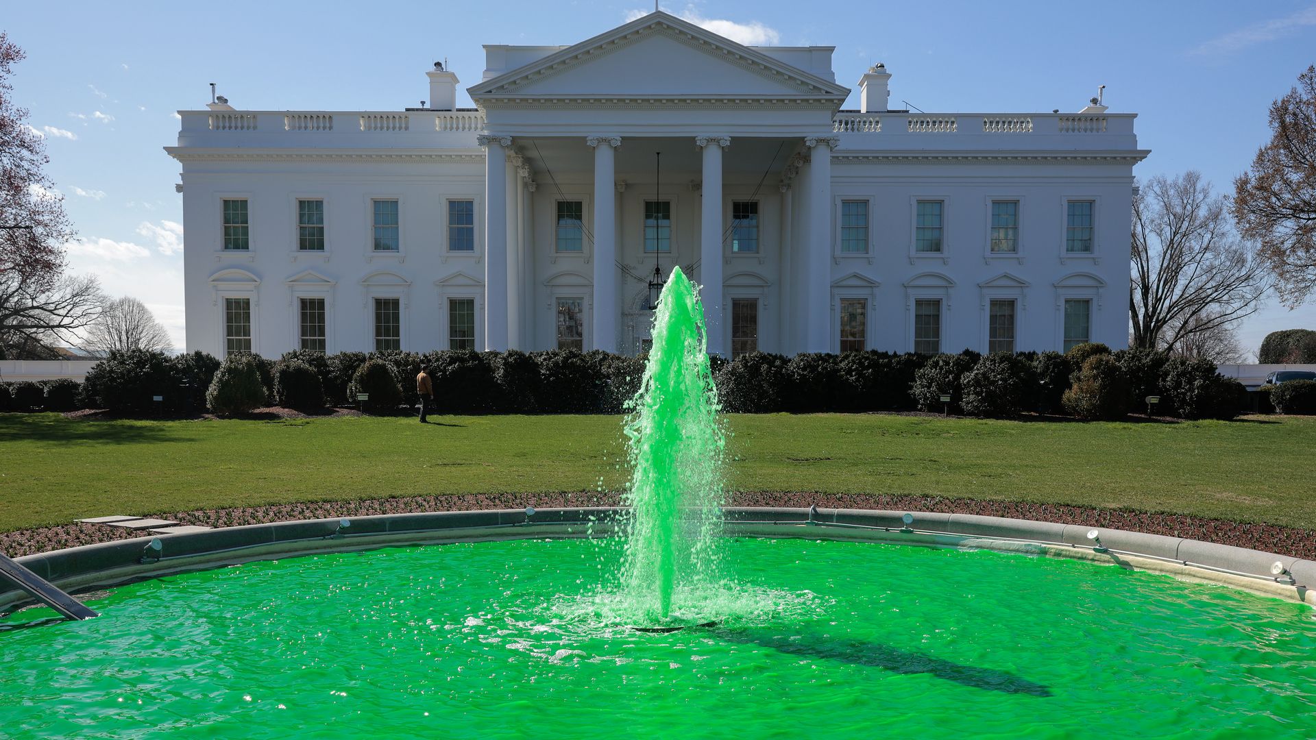 White neoclassical building, The White House, with tall columns and a triangular pediment, an American flag on top, a manicured lawn and hedges, and a bright green fountain spraying in front.