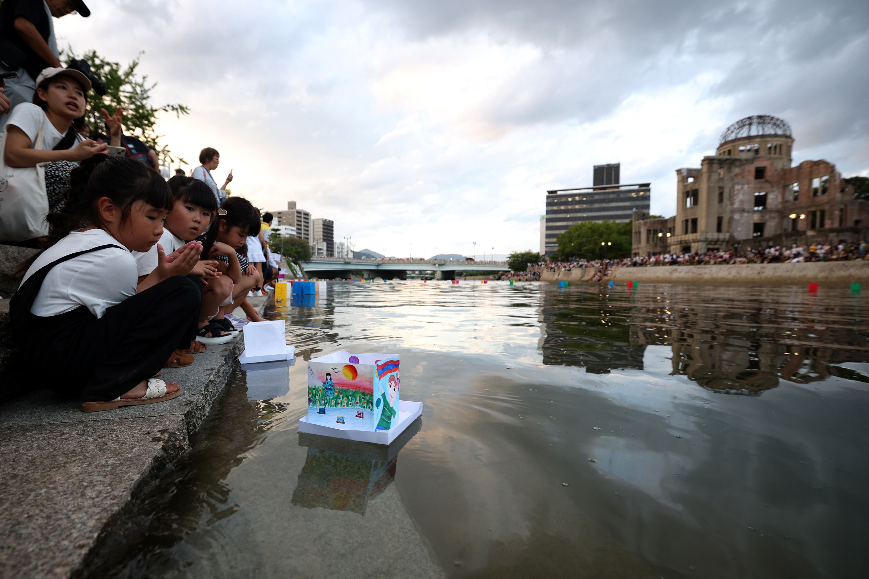 Children crouch at a river's edge releasing decorated paper lanterns on water during a solemn event, with a crowd and a historic dome-shaped ruin building visible under a cloudy sky.