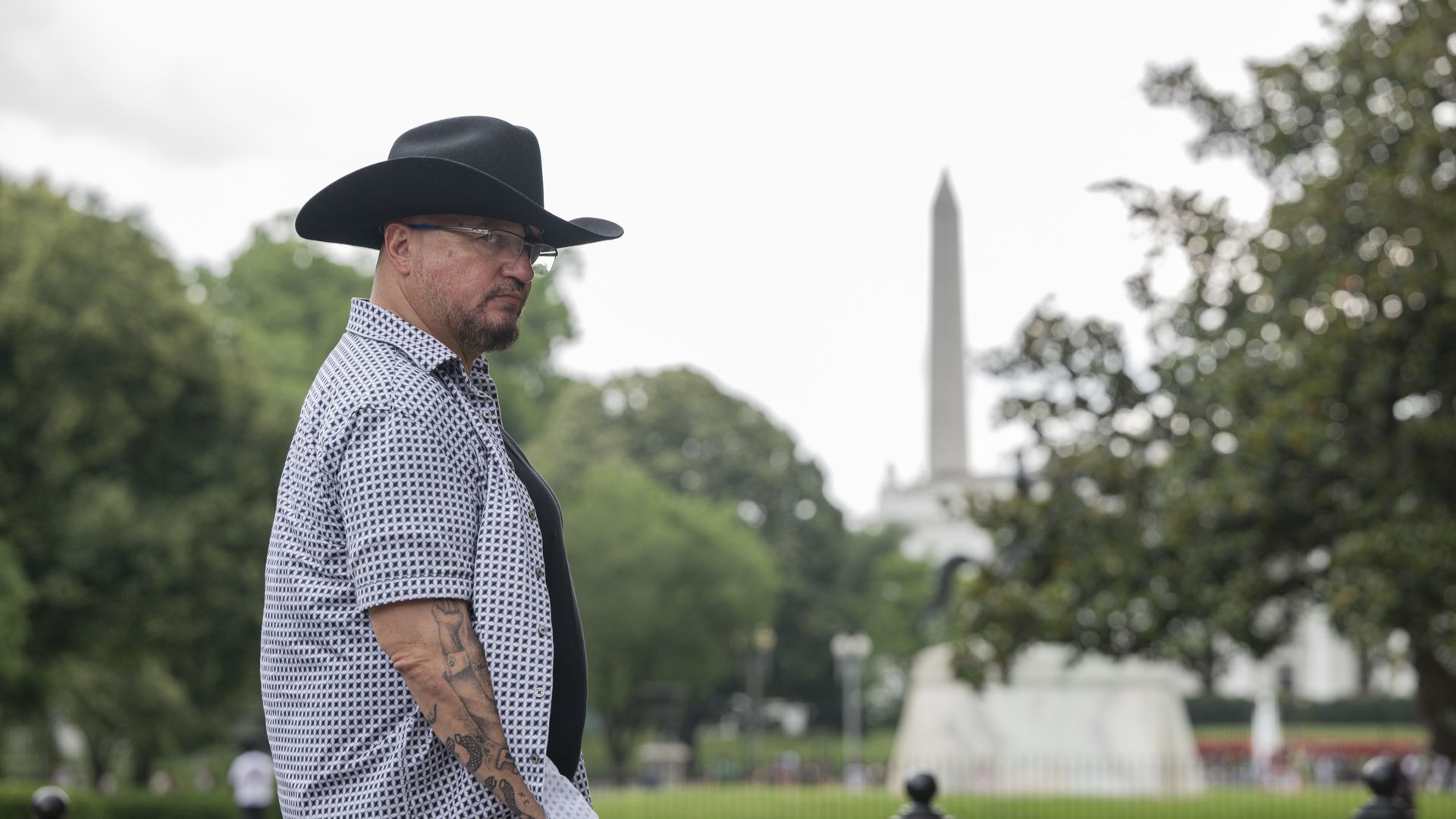 Stewart Rhodes stands to the side wearing a black wide-brimmed hat in front of the Washington Monument.