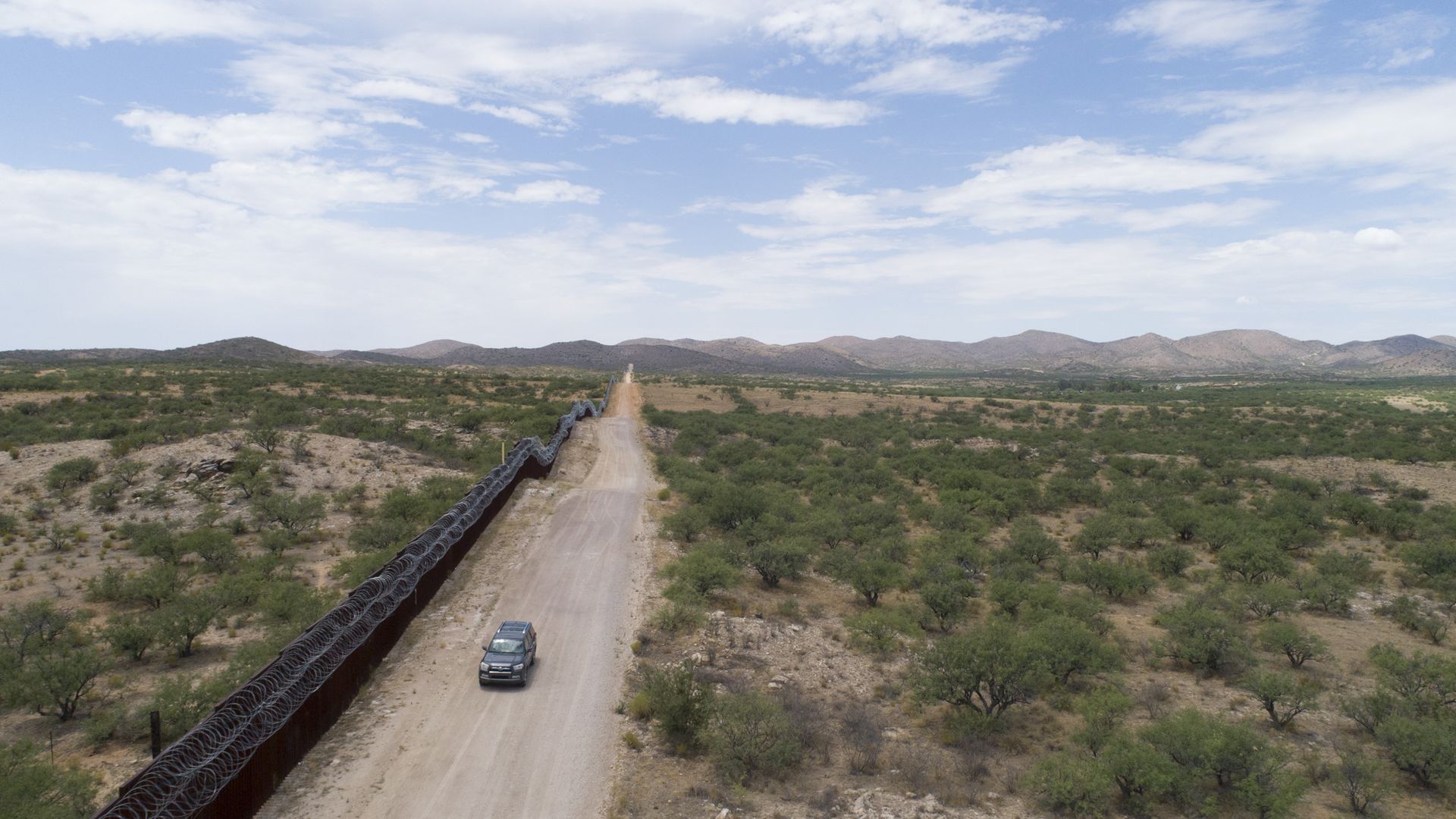 A car driving along a dirt road by a border fence on the southwest border.