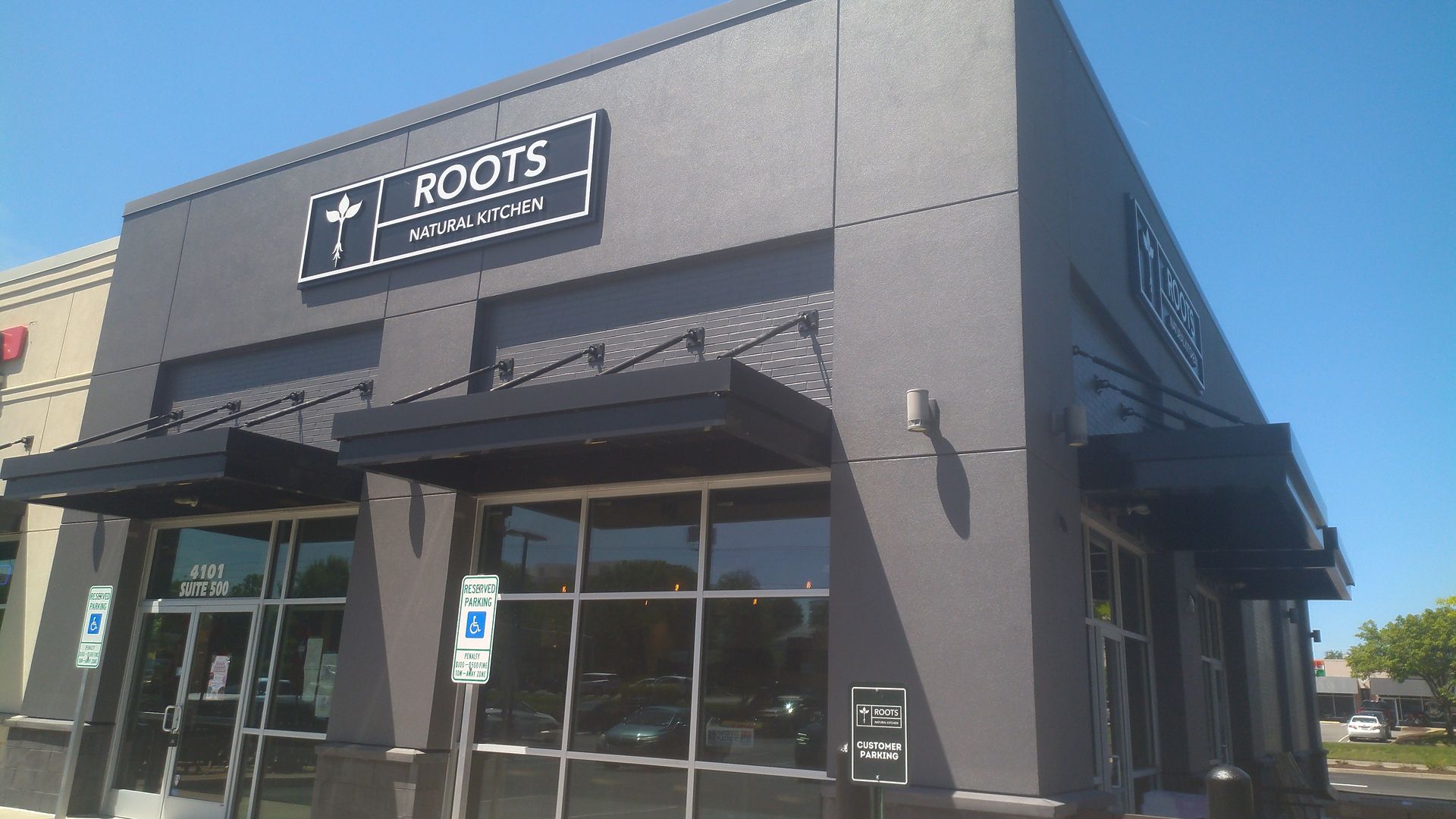 Roots Natural Kitchen storefront on a clear blue day; gray building, black sign with "ROOTS" and "NATURAL KITCHEN," plant logo, large glass doors.
