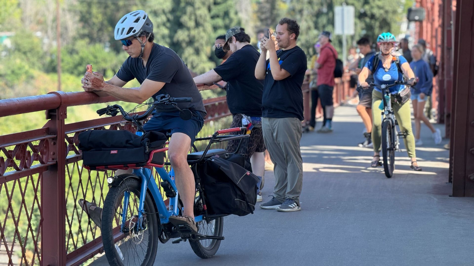 Group of cyclists on a red metal bridge under a blue sky. Foreground rider wearing a white helmet leans on railing with a bag; others ride behind, including a woman in a teal helmet.