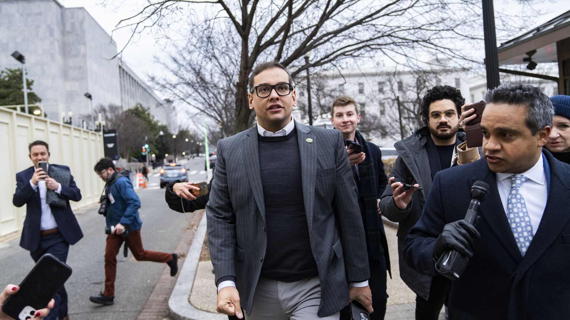 Rep. George Santos, wearing a gray pinstripe blazer, black sweater and white shirt, walks through the Capitol complex flanked by reporters.