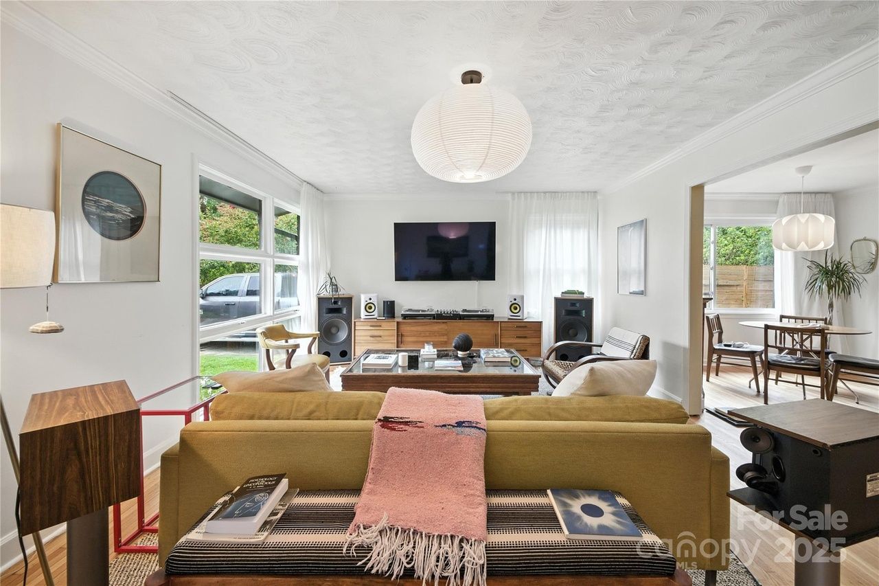 Bright, modern living room with white walls, a green sofa with pink throw blanket, wooden furniture, large windows, a flat-screen TV, speakers, and a view into a dining area with a round table and chairs.