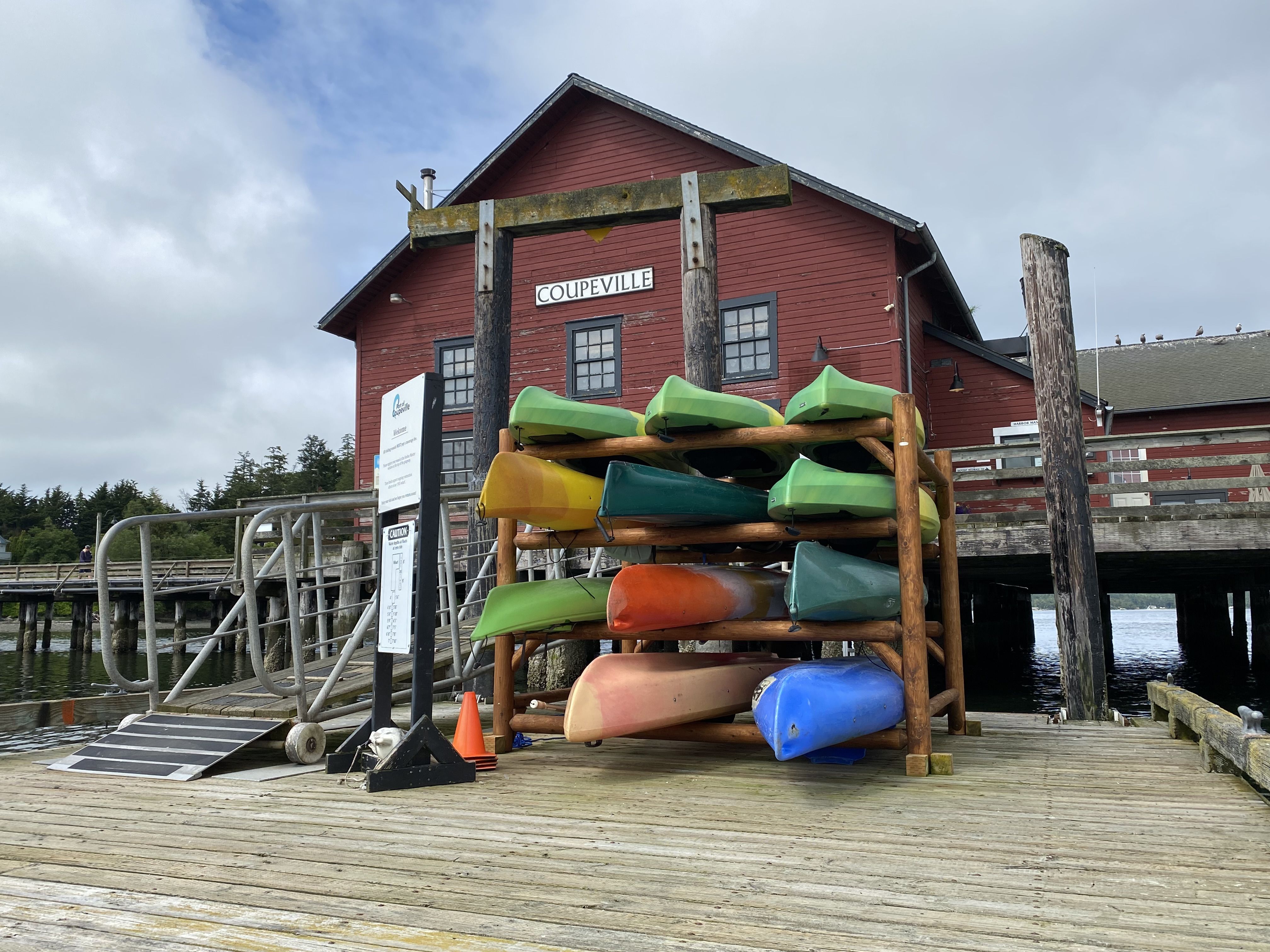 A red wharf building labeled "Coupeville" with colorful canoes and kayaks on a rack in front. 
