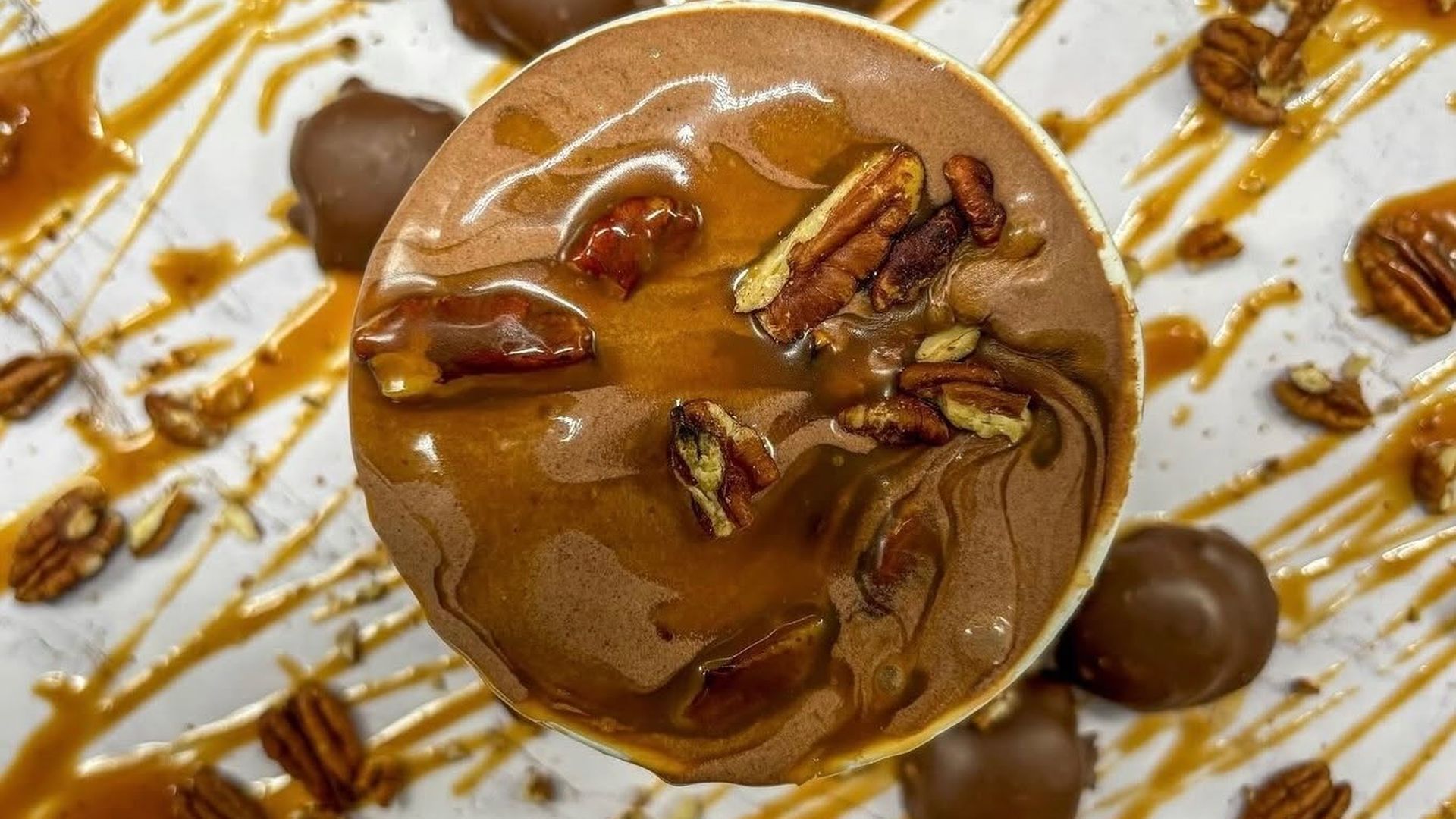 From above: a chocolate mousse cup topped with pecans and caramel sauce, surrounded by caramel drizzle, chocolate truffles, and scattered pecans on a white plate.