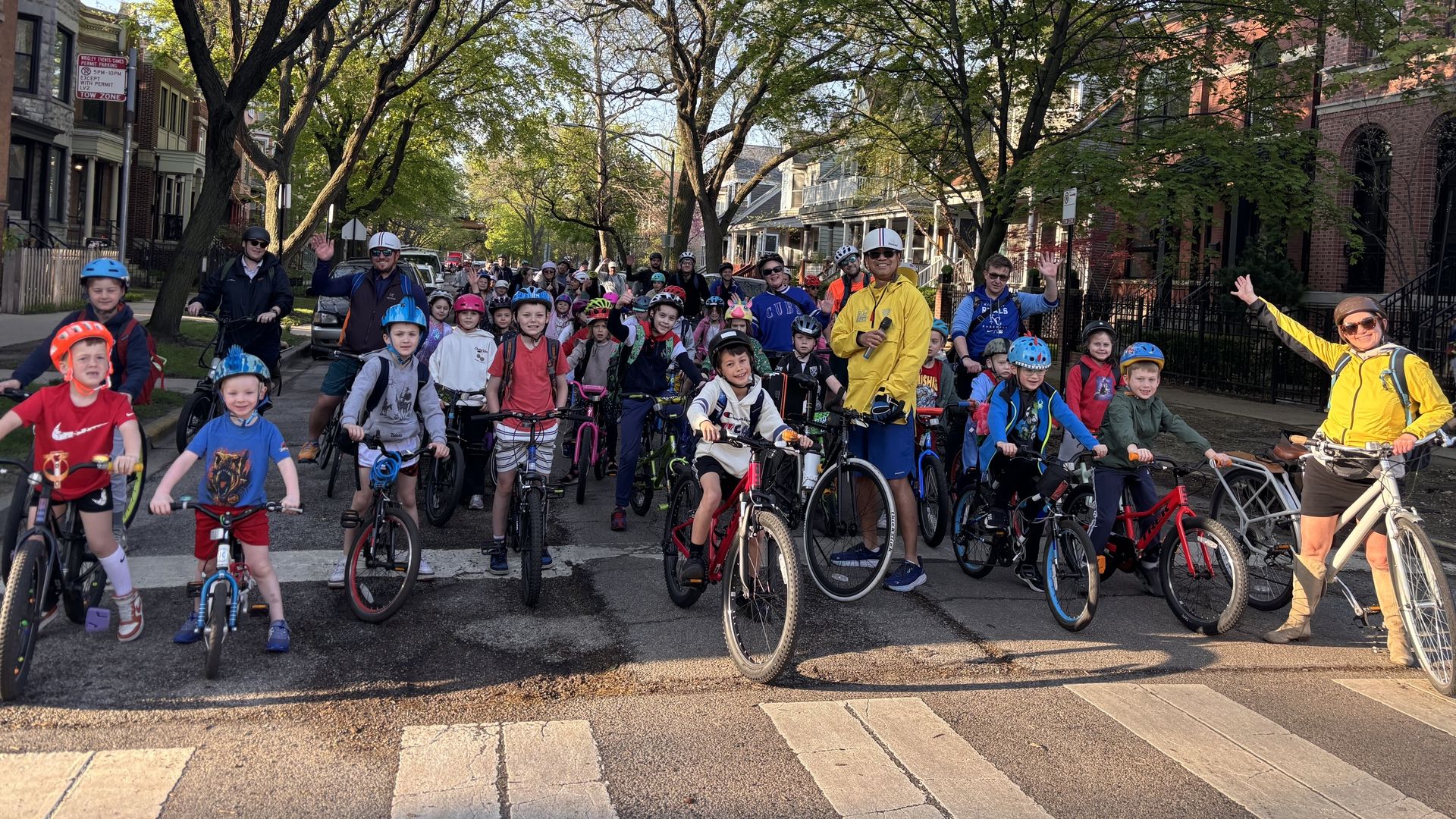 A crowd of children on bikes with helmets crowds a city street, guided by adults in bright jackets. Overhead, leafy trees arch between brick townhouses on both sides.