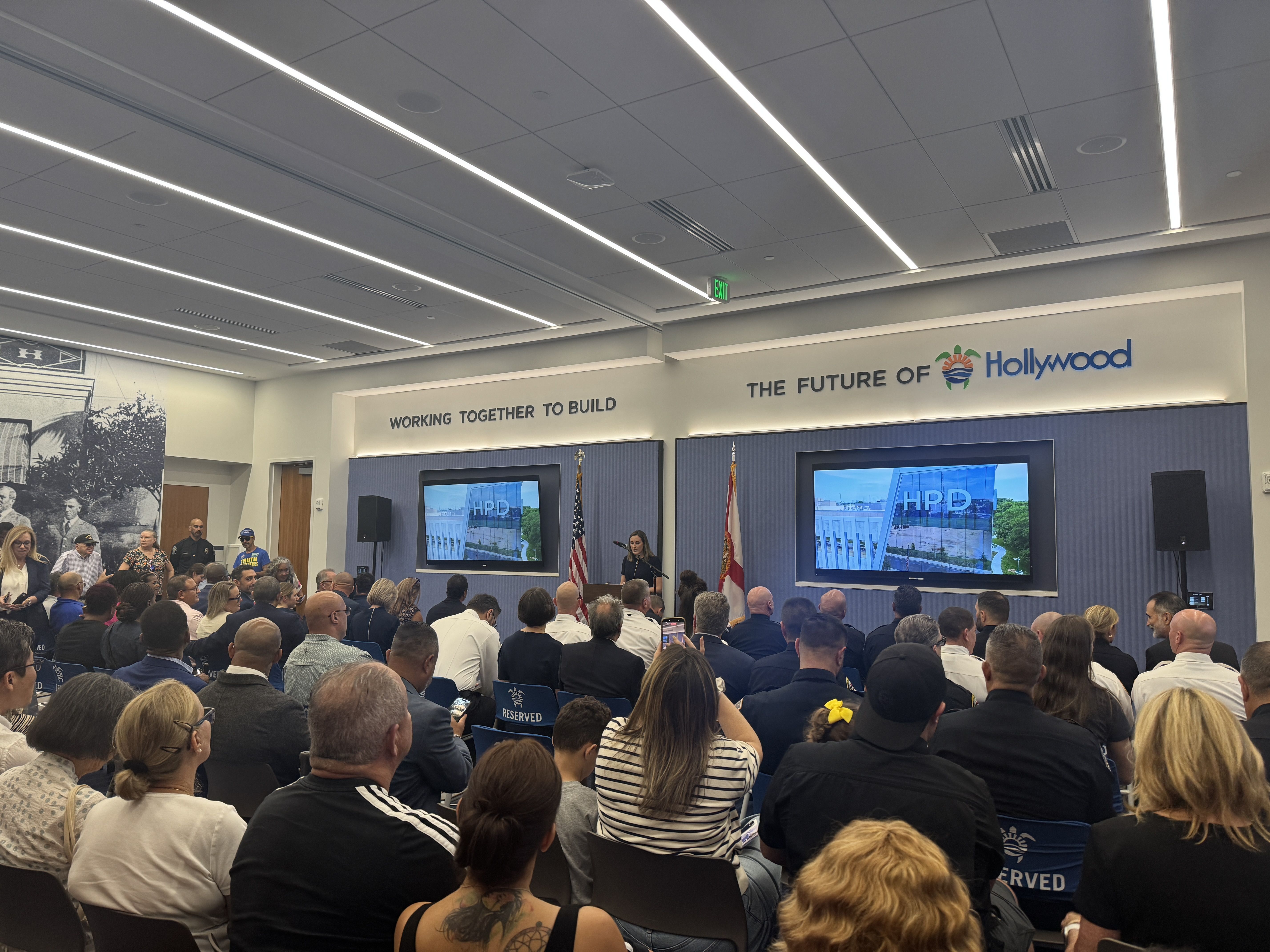 A large conference room filled with people facing a stage labeled "The Future of Hollywood" with screens showing a building, American flags, and a speaker at a podium.