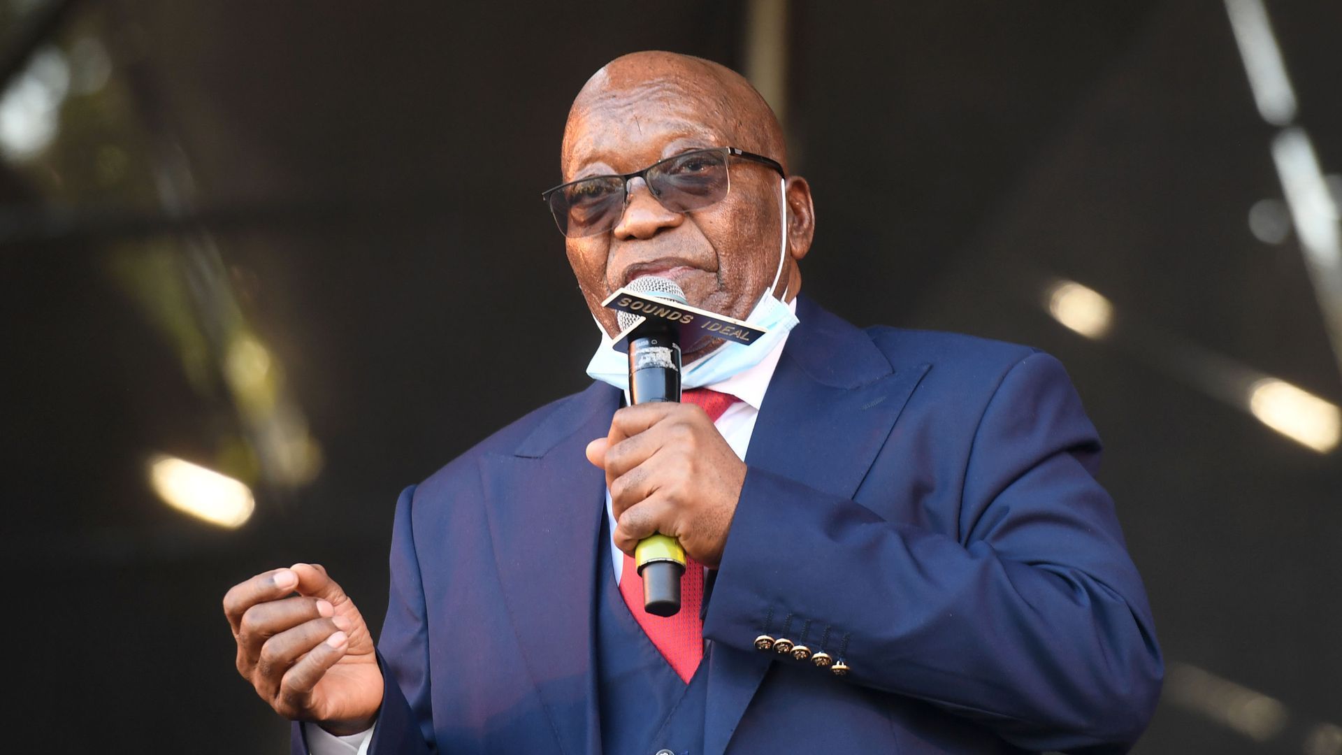 Former South African President Jacob Zuma, in a navy blue suit and red tie, speaks into a microphone as he addresses supporters.