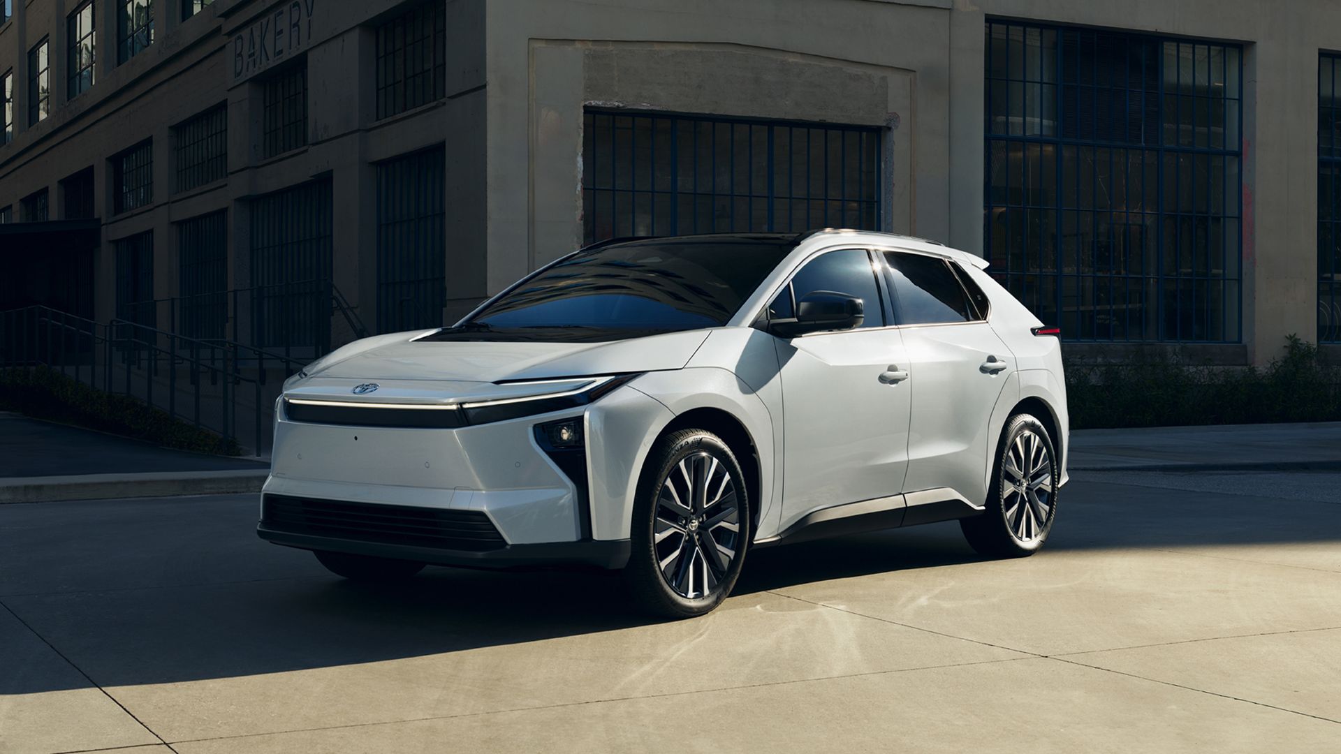 White Toyota bZ electric SUV with dark windows sits parked on a city street in front of a gray building with blue-framed windows; sun glints off the roof rails.