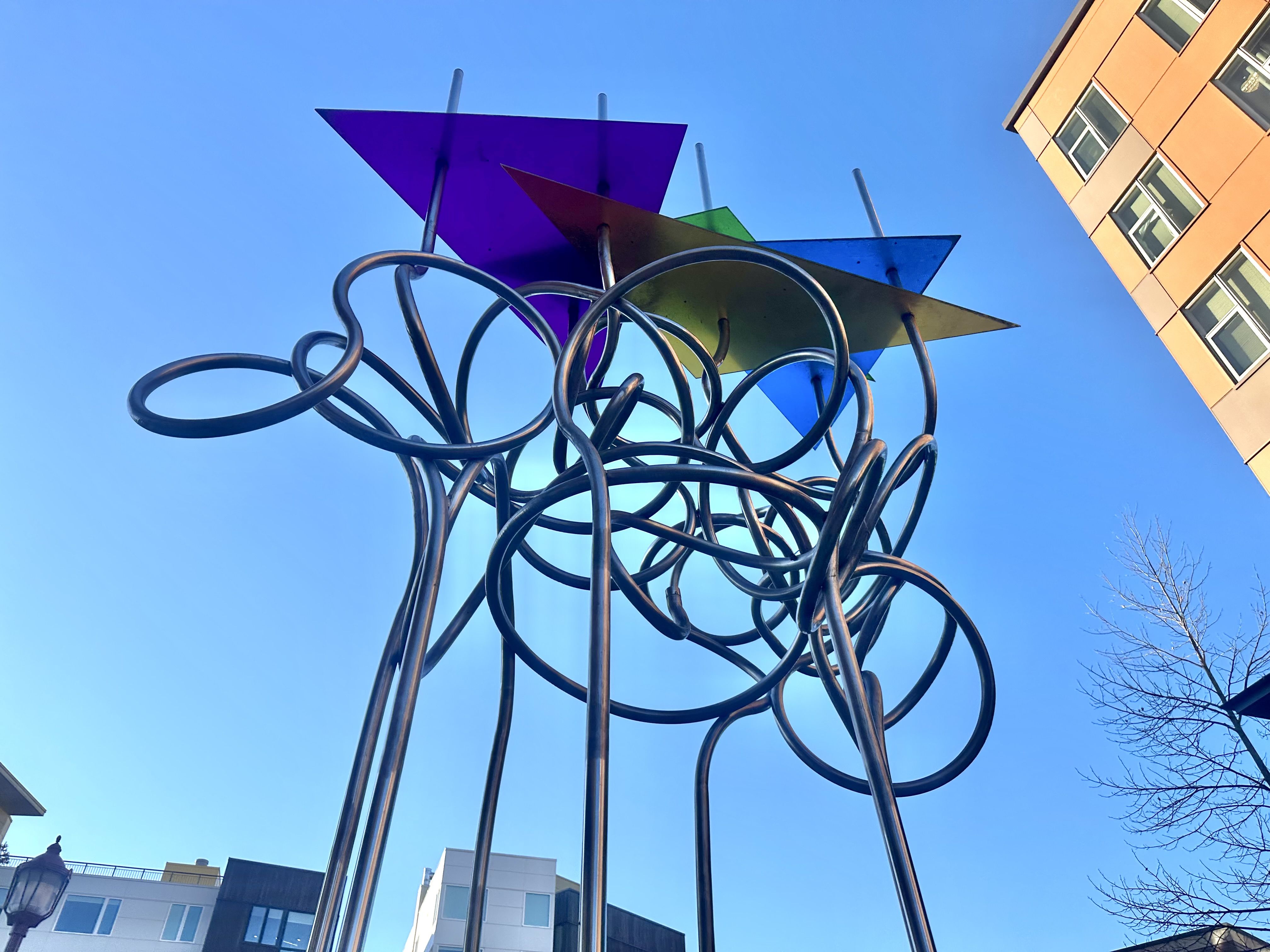 Metal poles rise and twist into squiggles, with colorful triangles on top and buildings to the side.