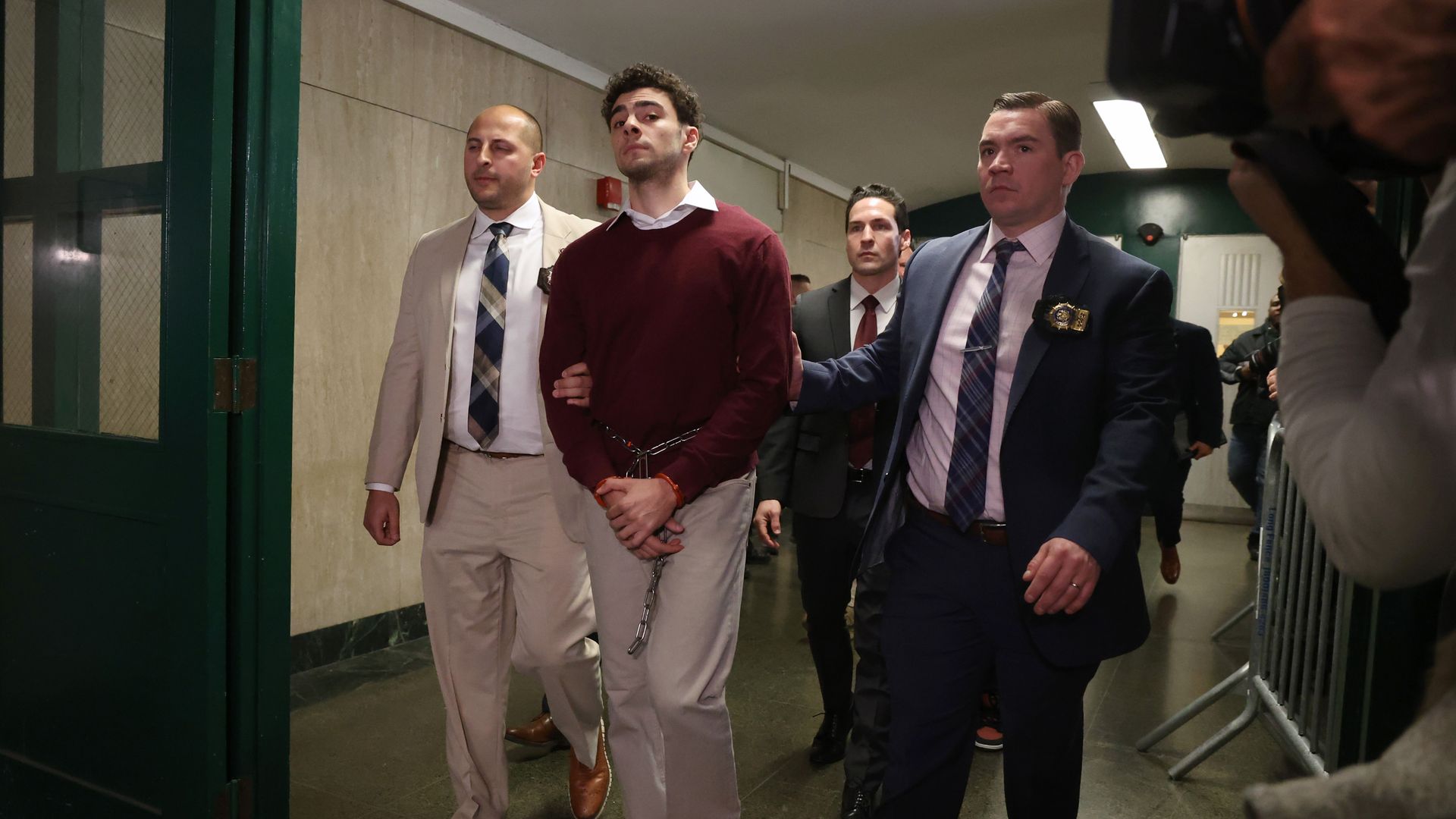 Luigi Mangione, wearing a burgundy sweater and khaki pants, is flanked by people guiding him through a hallway. His hands are restrained.