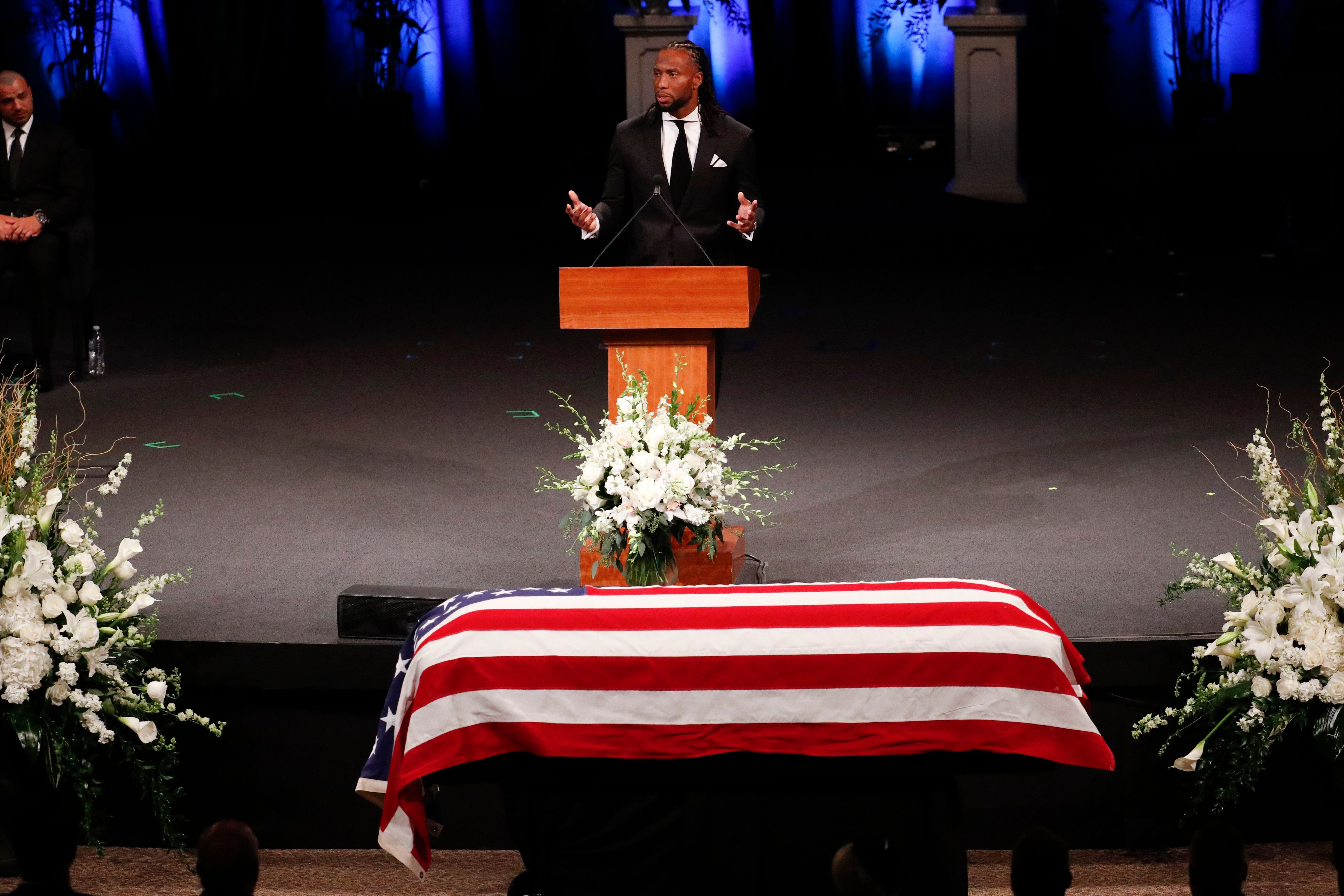 A man speaking in front of a casket draped with an American flag.
