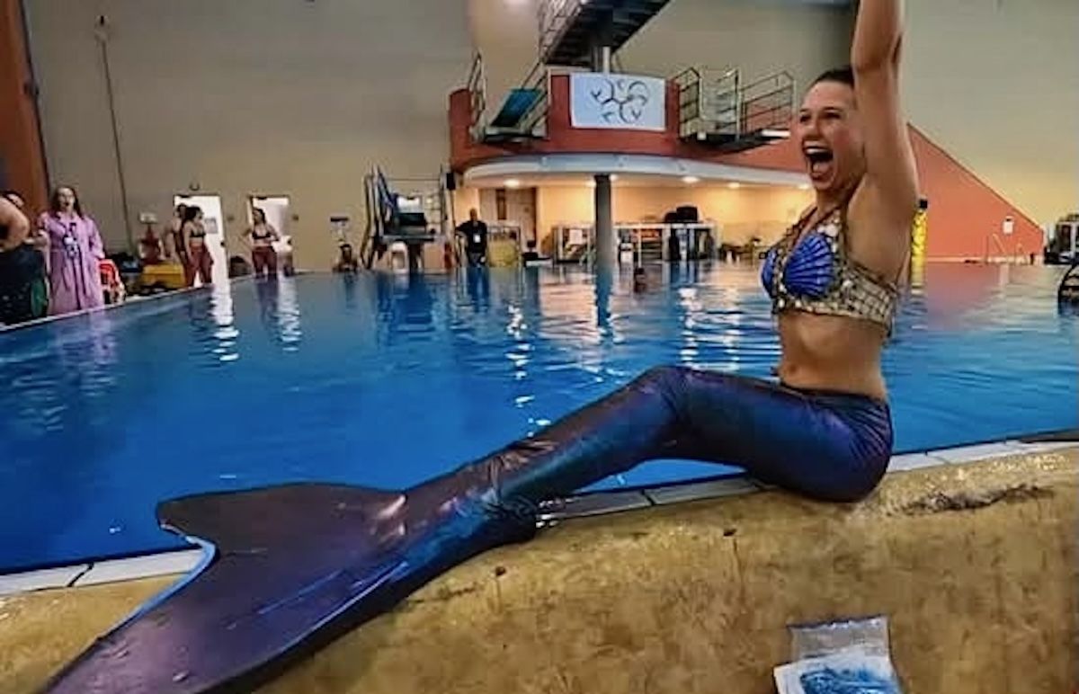 A woman dressed as a mermaid jubilantly waves her arms next to a swimming pool.