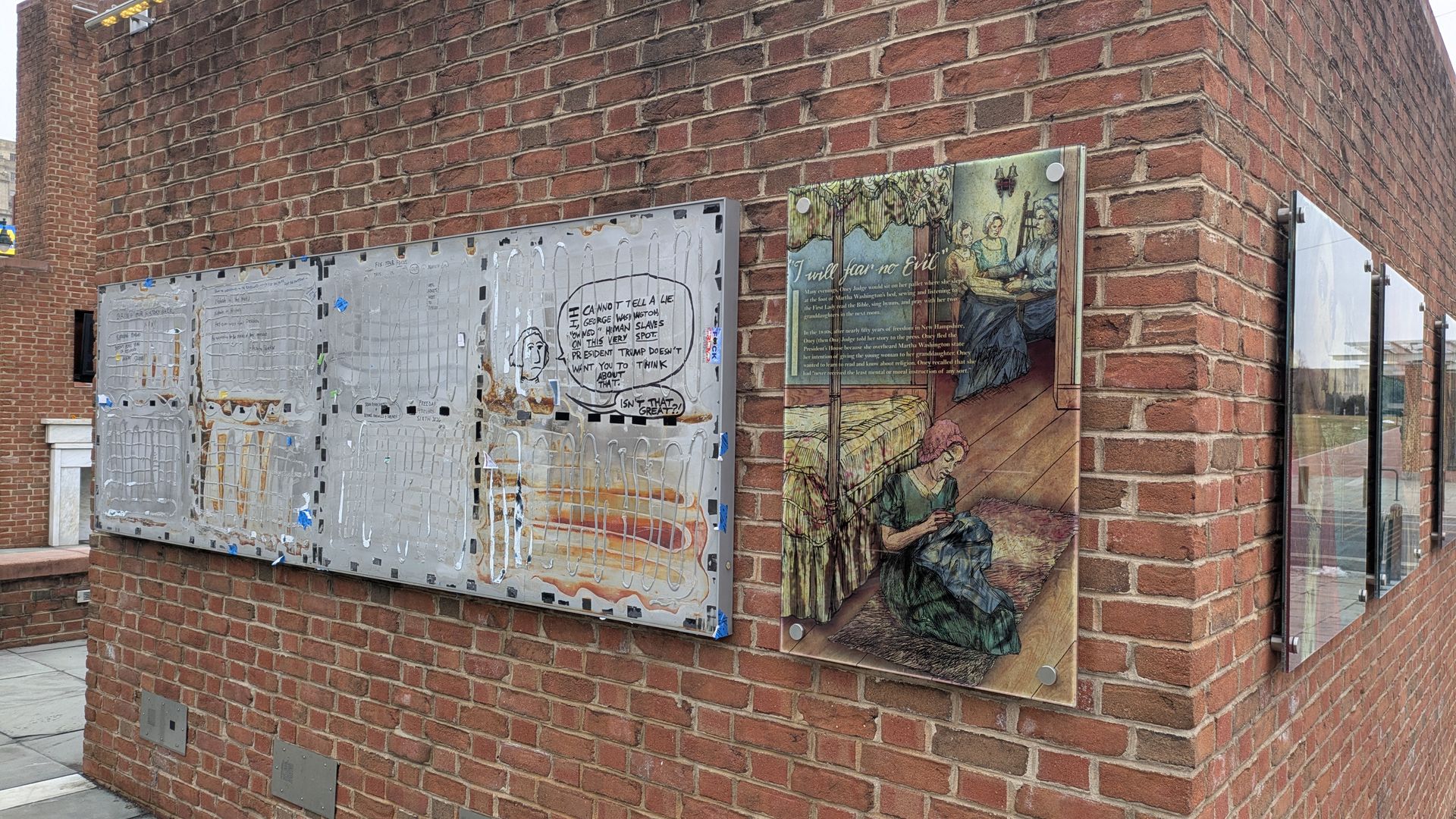 Brick wall corner with multiple plaques; one plaque has a colored historical illustration of a woman sewing on a rug with text titled "I will fear no Evil"; another metal plaque has worn text and a speech bubble illustration.