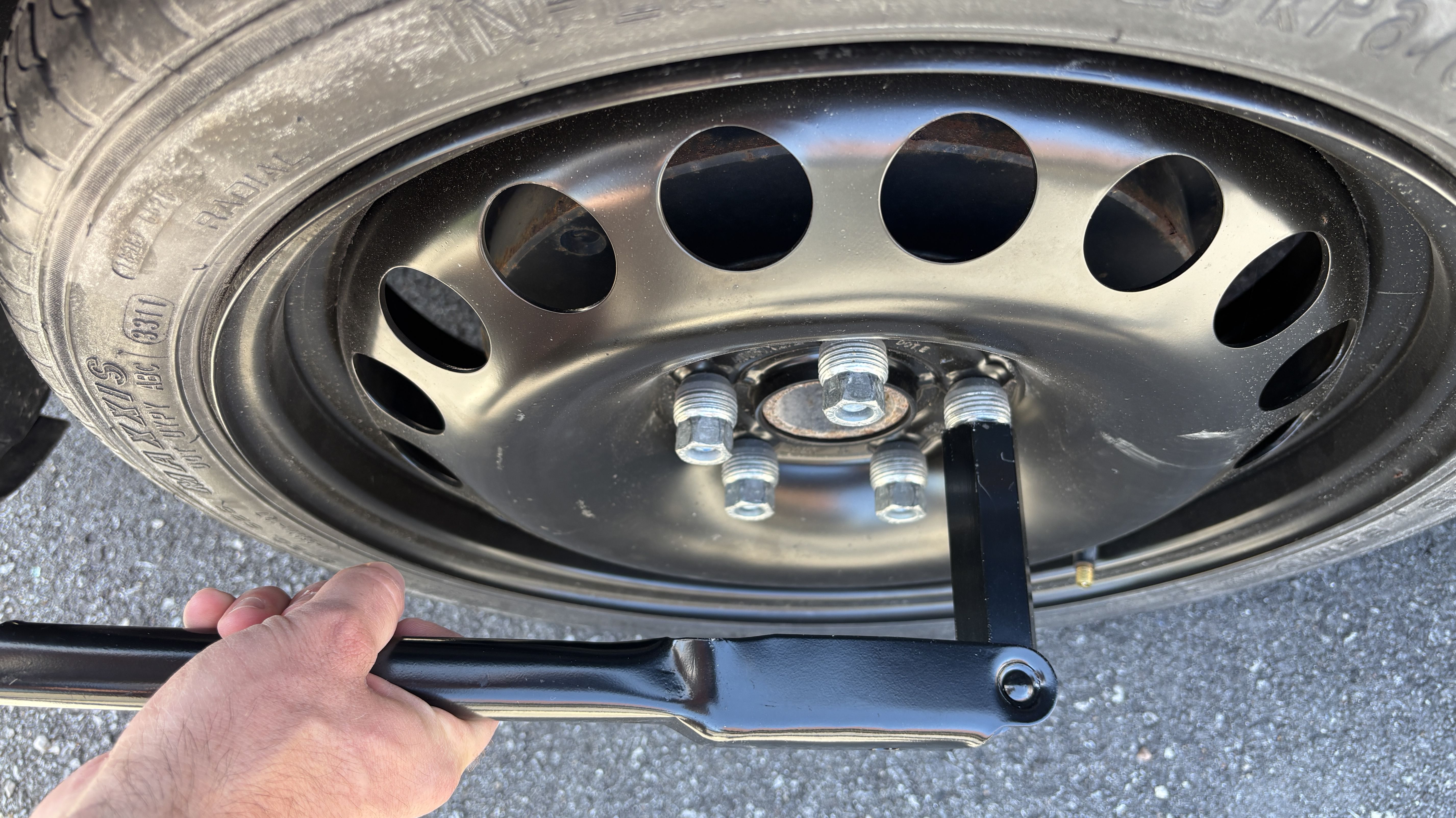 Close-up of a car wheel with a black steel rim featuring circular cutouts, five lug nuts, and a scissor jack nearby; a hand is visible at the lower left.