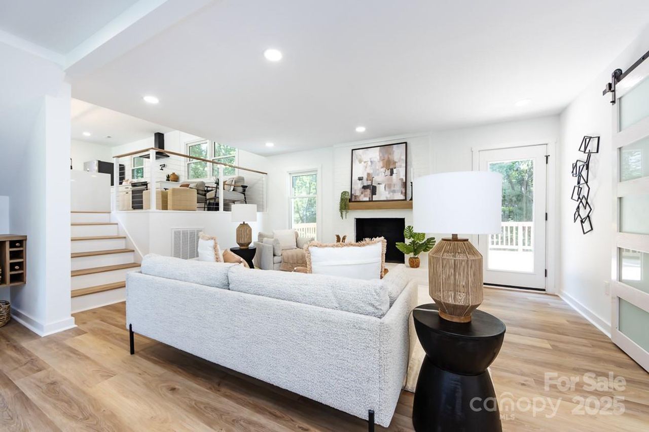 Bright living room with light gray sofa, wooden floor, black side table with lamp, abstract painting above fireplace, stairs leading to an elevated dining area with large windows.