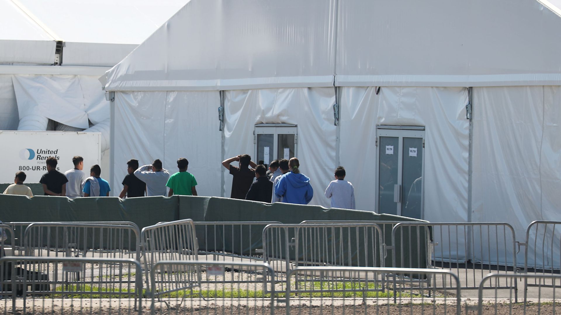 Children walk in a line out of a large white tent with double doors at the Homestead facility. 