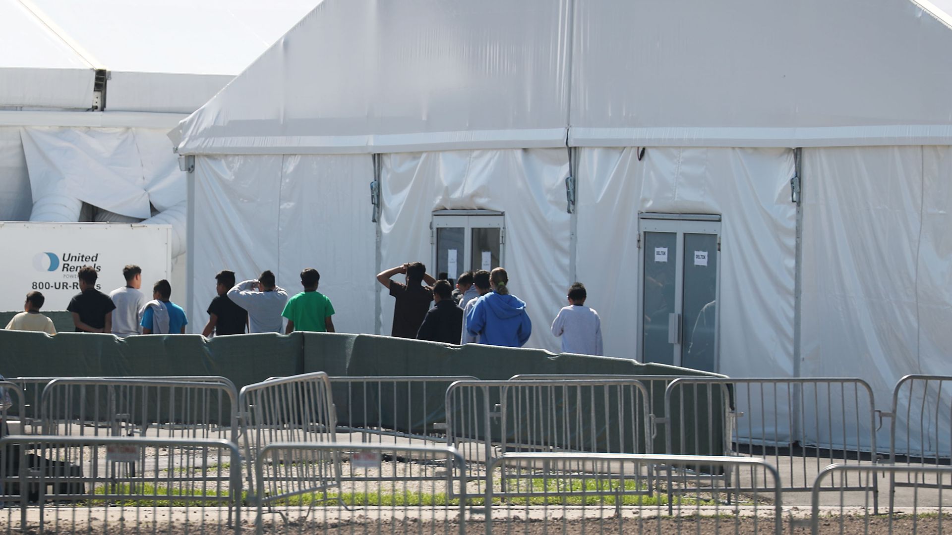 Children walk in a line out of a large white tent with double doors at the Homestead facility.