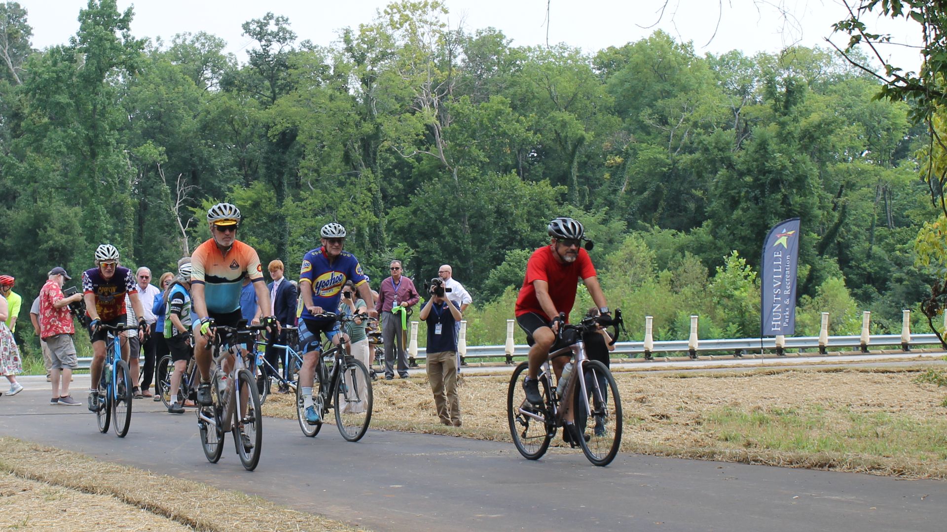 Cyclists in colorful jerseys ride a paved path at a race event; spectators line the roadside as trees and greenery form a lush background.