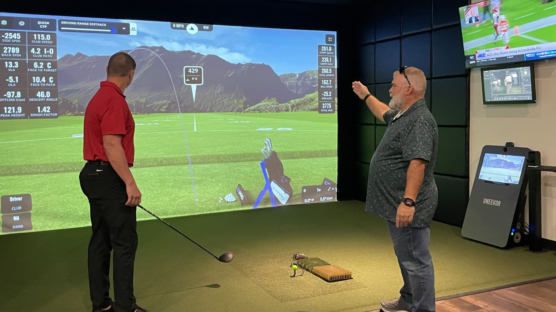 Two men playing indoor golf using a golf simulator projecting a mountain scene and ball trajectory; one holds a club preparing to swing, the other gestures while explaining. TVs on wall.