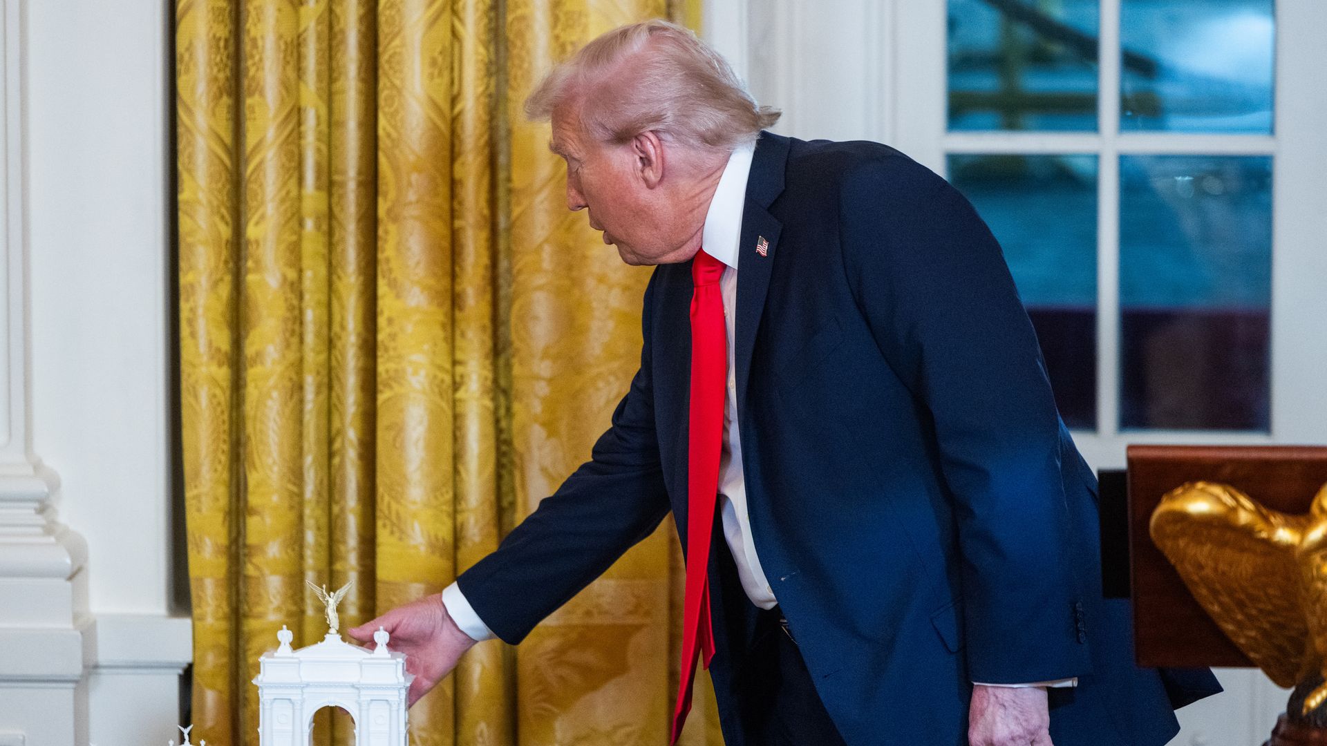President Trump — wearing a dark blue suit, a white collared shirt, a red tie and an American flag pin — reaches for a small, white model of an arch on a table.